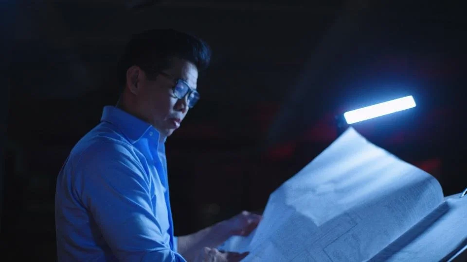 A man wearing glasses and a blue shirt reading a large book in a dimly lit room with a bright light illuminating the pages.