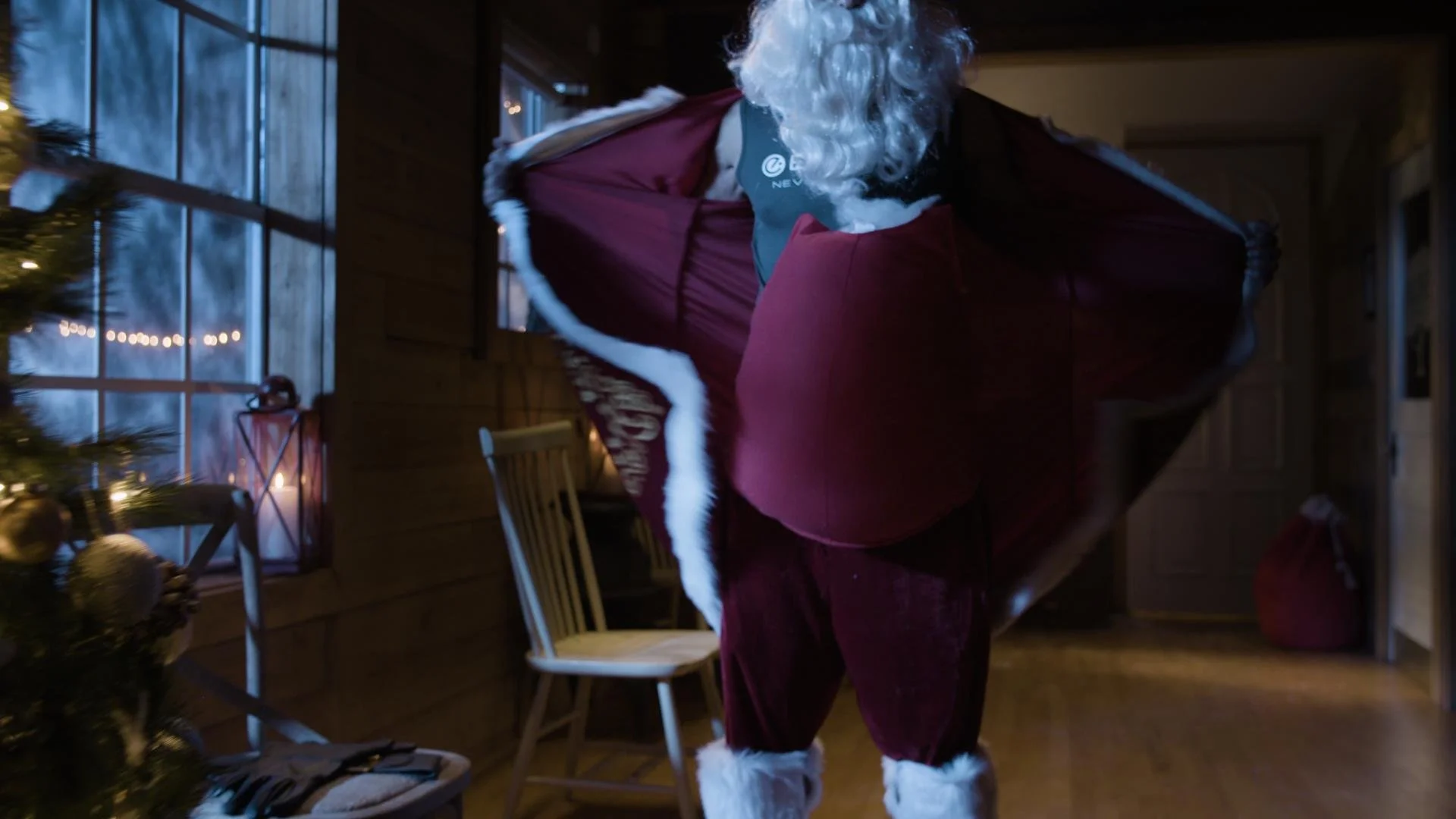 Santa Claus in a red suit and hat, with white fur trim, carrying a large gift box inside a cozy, decorated home with a Christmas tree, window, and wooden walls.