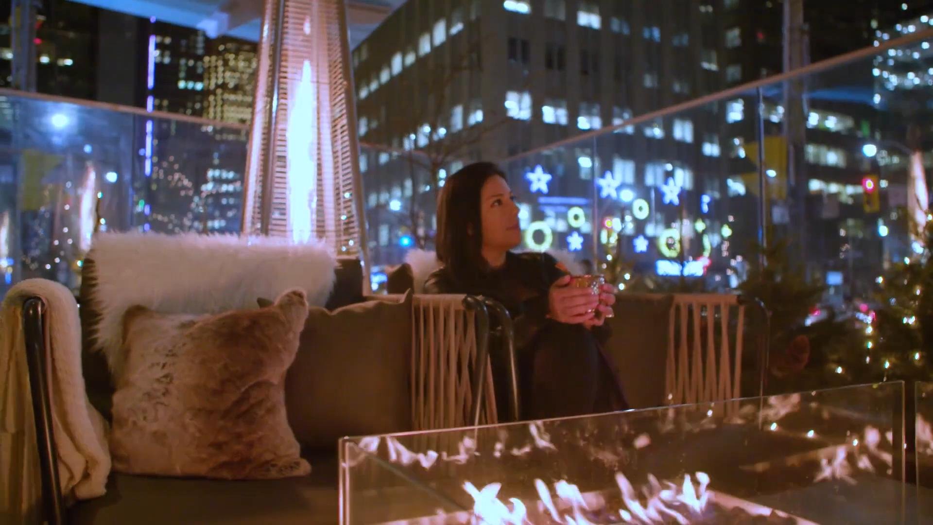 A woman sitting on a balcony at night, holding a mug, with Christmas lights and decorations in the background, and a cat on a couch nearby.
