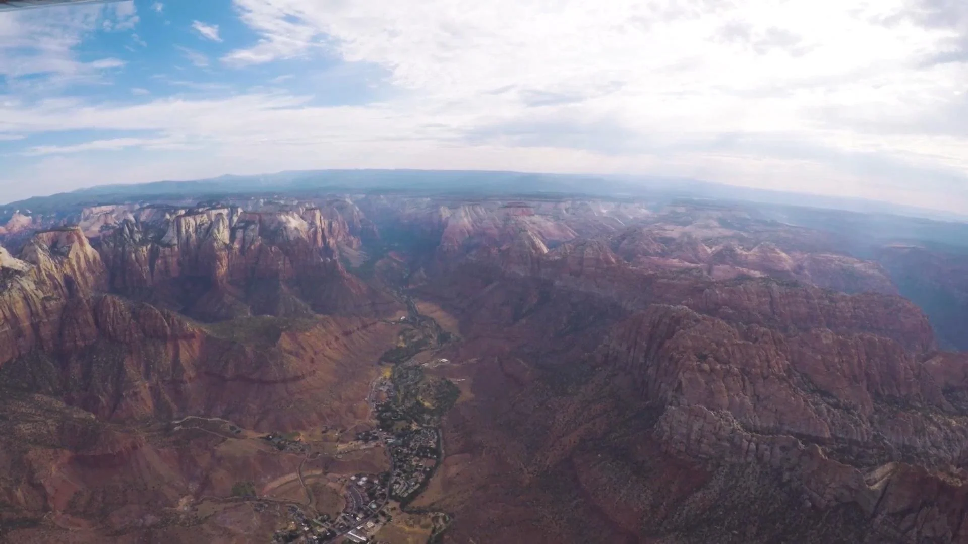 Aerial view of the Grand Canyon with layered red and brown rock formations, a river running through the bottom, and a partly cloudy sky above.