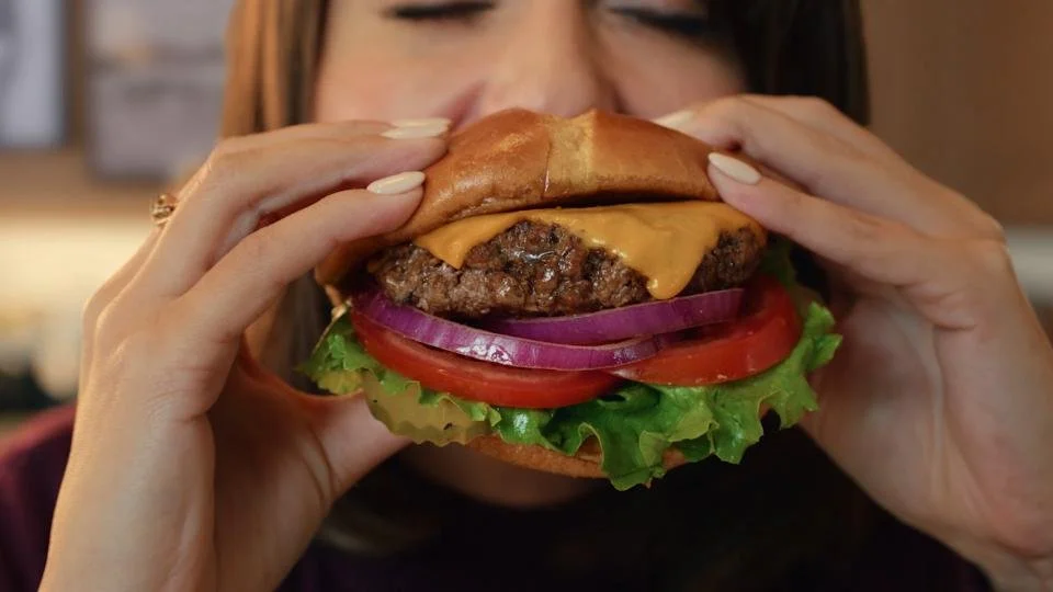 Person holding a cheeseburger with lettuce, tomato, onions, and melted cheese.