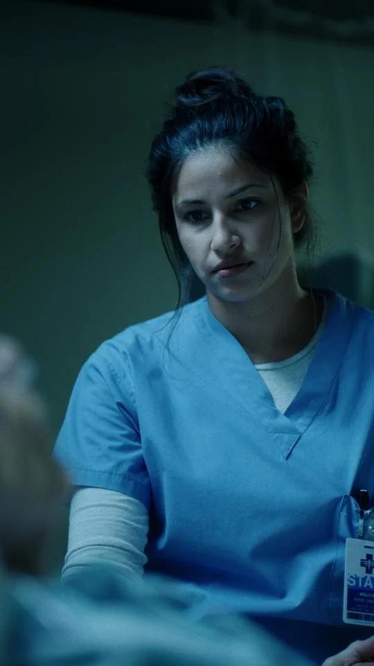 A young woman in blue medical scrubs and a white shirt looks intently at someone or something off-camera, standing in a dimly lit room.