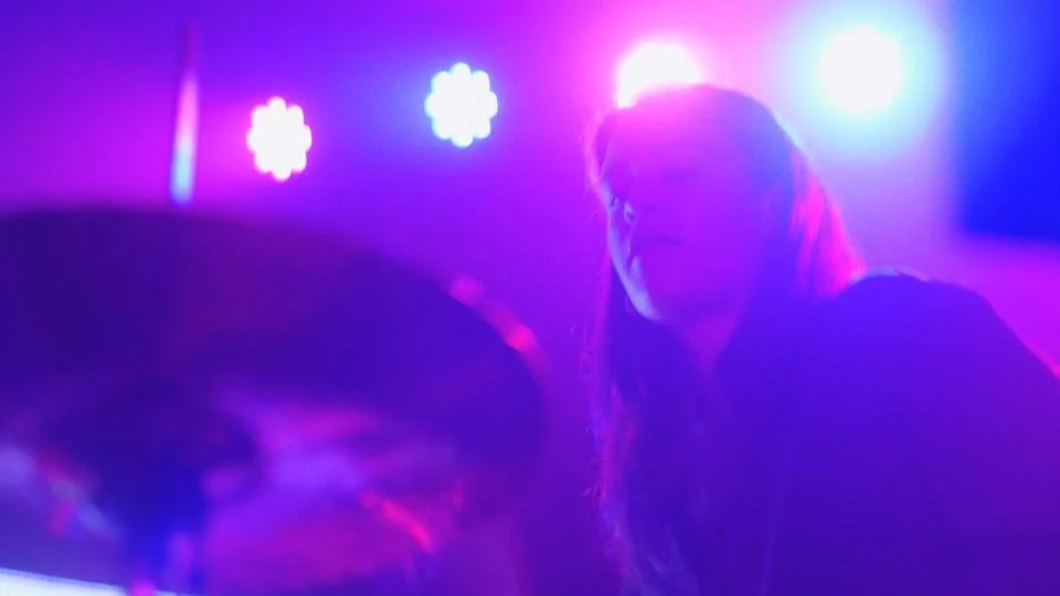 A woman with long hair in a dark room with colorful purple and pink stage lighting.