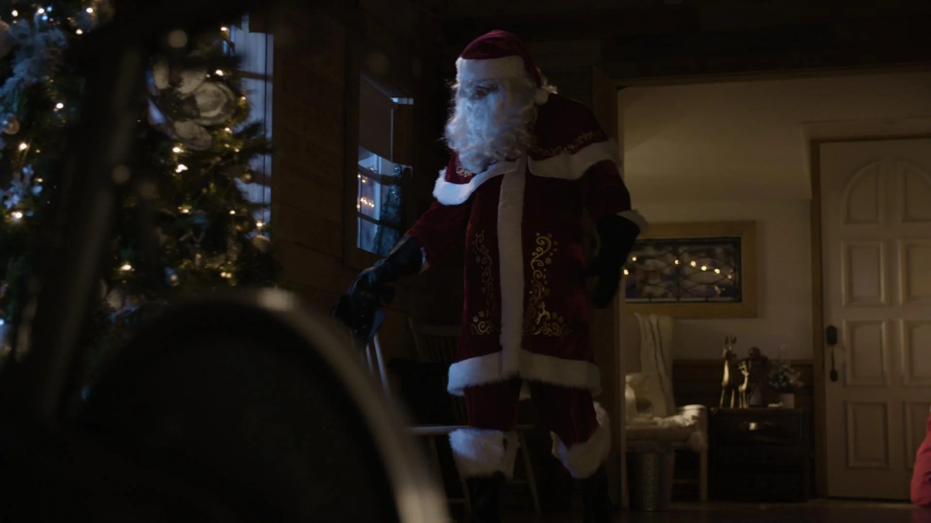 A person dressed as Santa Claus standing indoors near a decorated Christmas tree, with warm lighting and holiday decorations in the background.