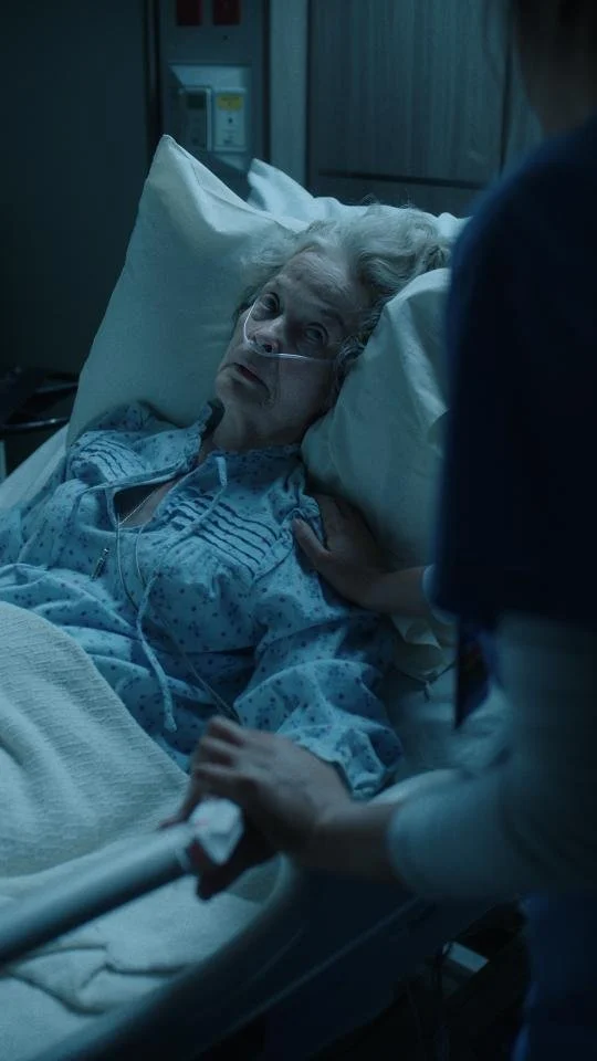 An elderly woman lying in a hospital bed with oxygen tubing, looking at a healthcare worker beside her.