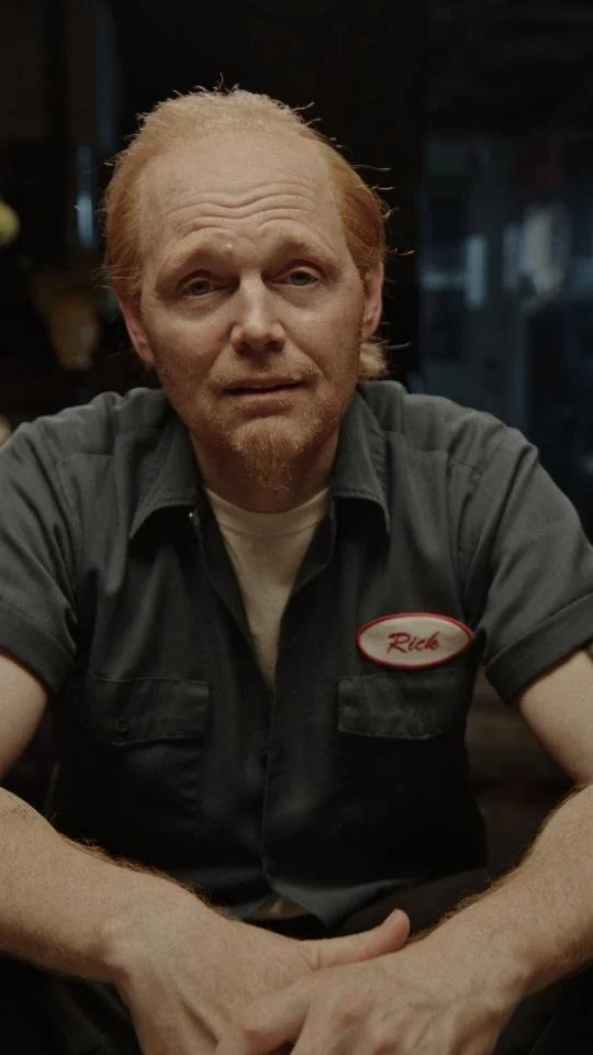 A man with red hair and beard wearing a dark gray work shirt with a name tag that reads "Rich" sits at a table in a dimly lit restaurant.