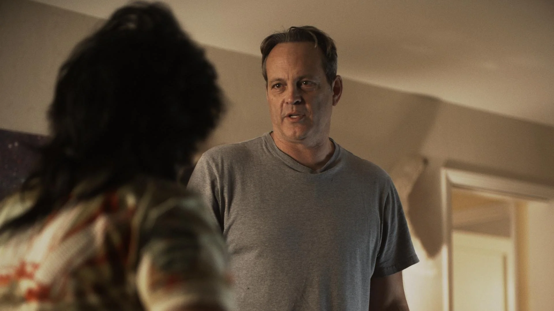 A man with short hair wearing a gray t-shirt is talking to a person with dark, curly hair and a patterned shirt, in a room with beige walls and an air conditioning unit in the background.