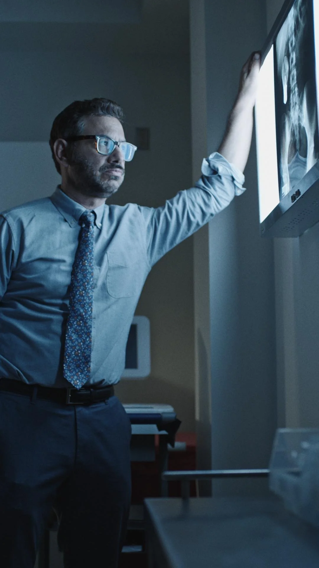 A man in a shirt and tie, wearing glasses, examining an X-ray on a lightbox in a dimly-lit room.
