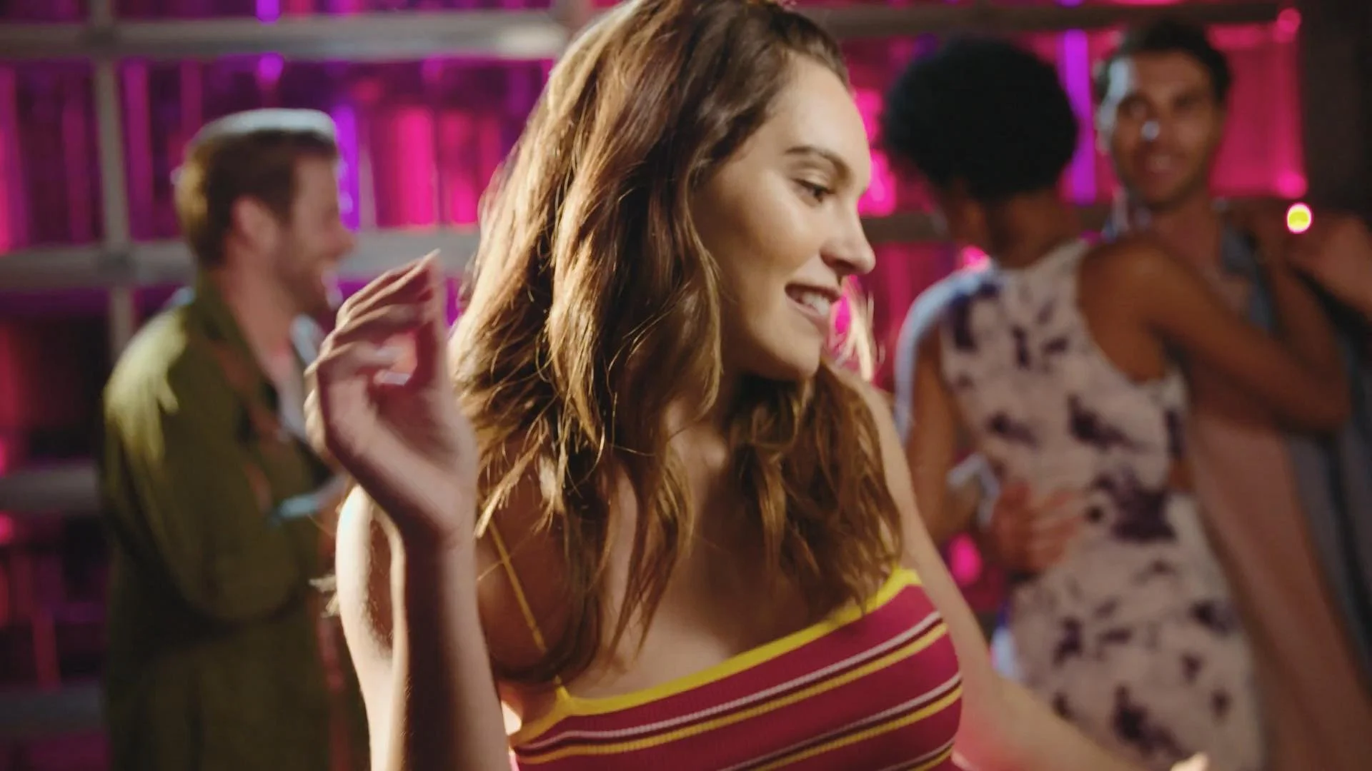 A young woman dancing at a lively indoor party with colorful lighting, smiling with eyes closed, wearing a striped yellow and red top, surrounded by other people enjoying the music.