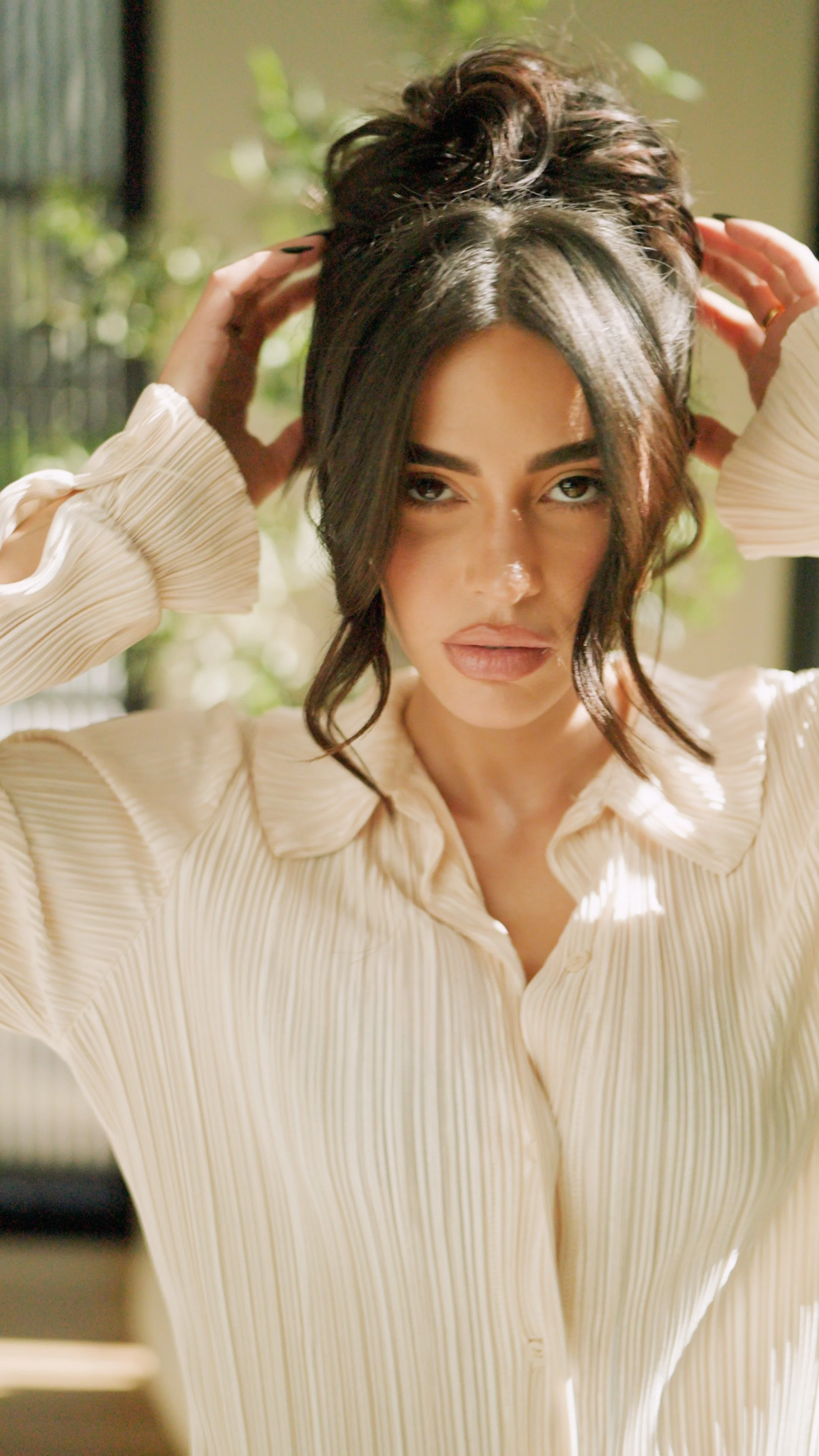 A woman with long dark hair, wearing a cream-colored, pleated blouse, styling her hair in a bright room with natural lighting and a blurred background.