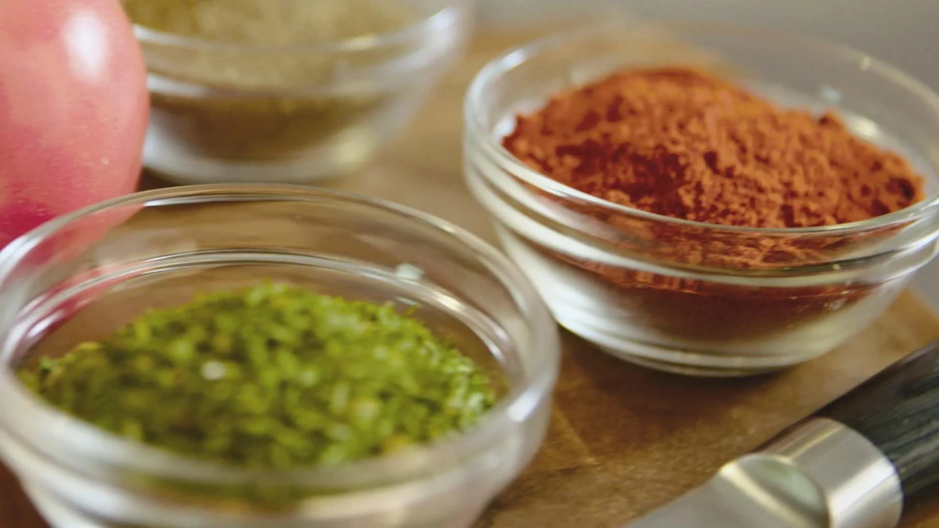 Close-up of small glass bowls containing various spices and herbs on a wooden surface, with an apple partially visible on the left side.