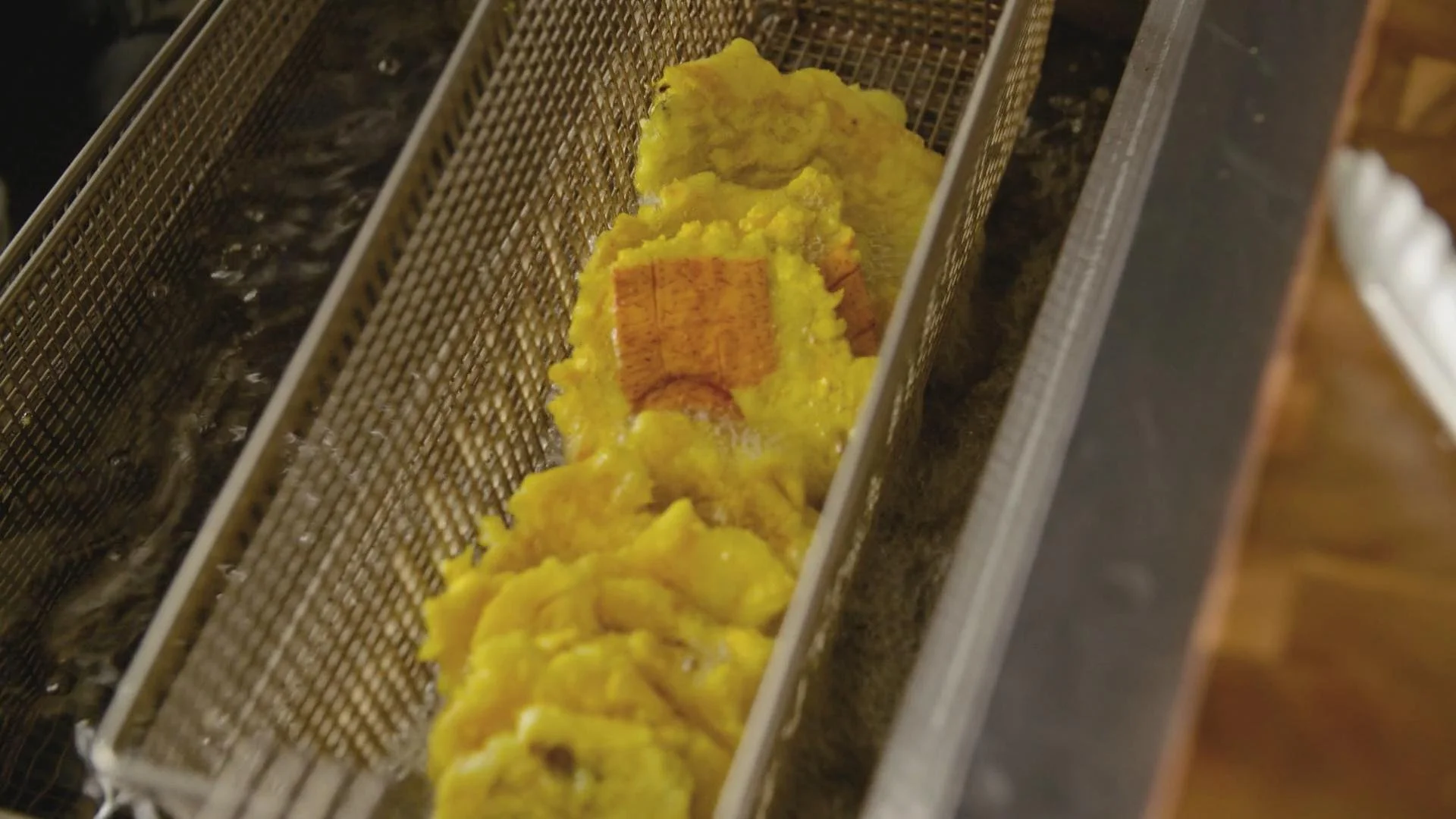 Yellow battered food being fried in a metal mesh basket at a deep fryer.
