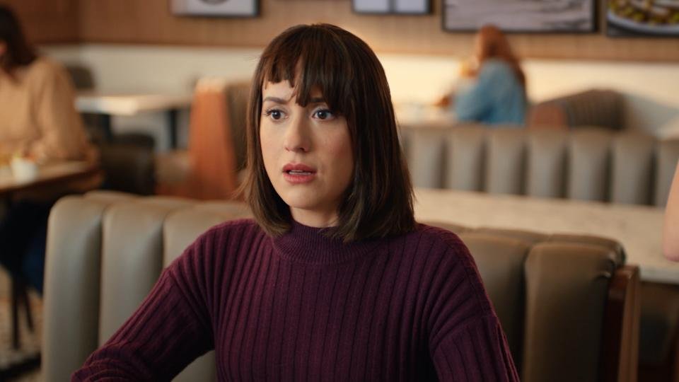 A woman with shoulder-length brown hair and bangs, wearing a maroon ribbed sweater, sitting in a restaurant with a surprised or confused expression.