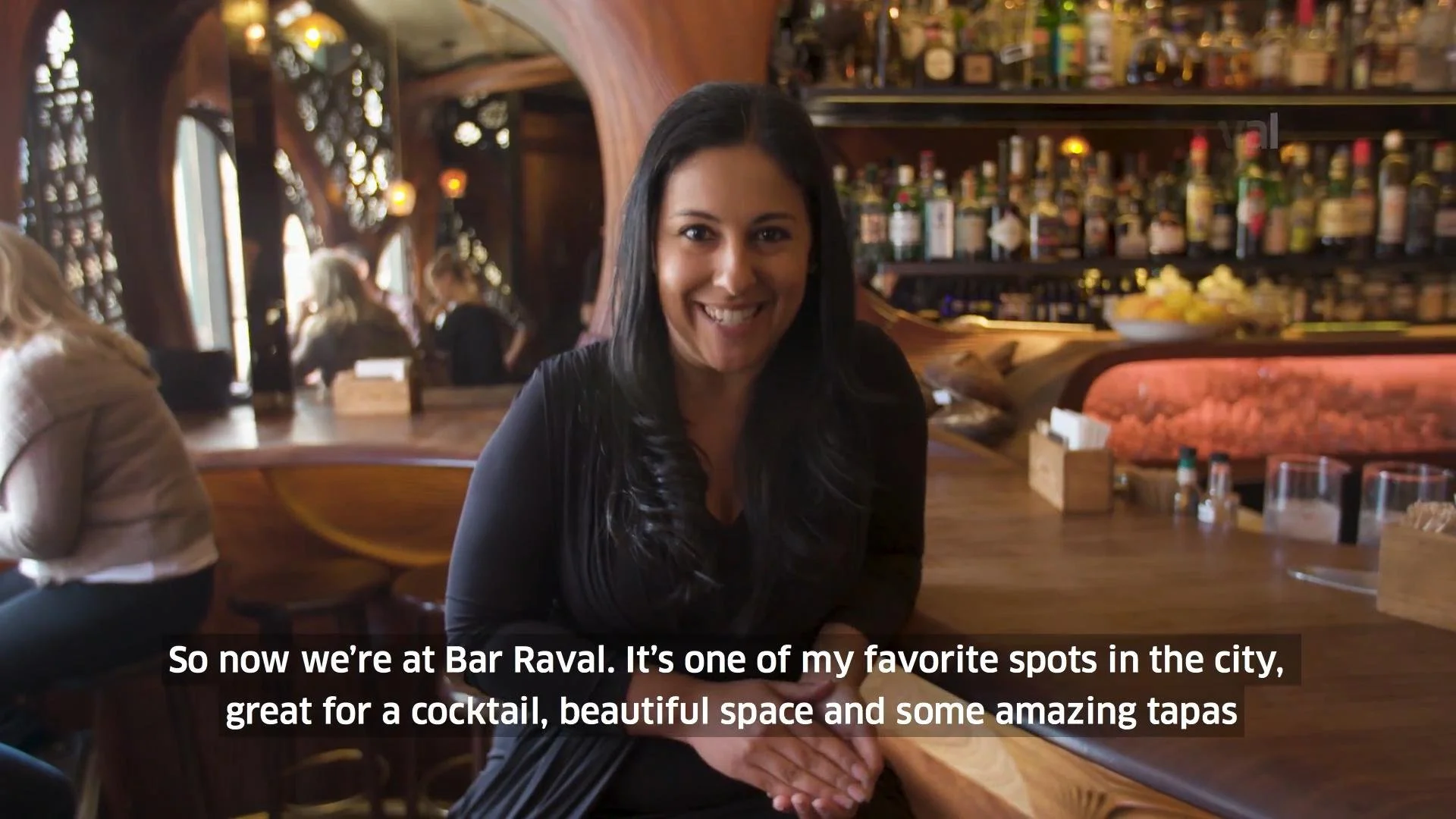 A young woman with dark hair and a black top sitting at a wooden bar in a warmly lit restaurant, smiling at the camera. The background shows shelves with bottles and a cozy ambiance.