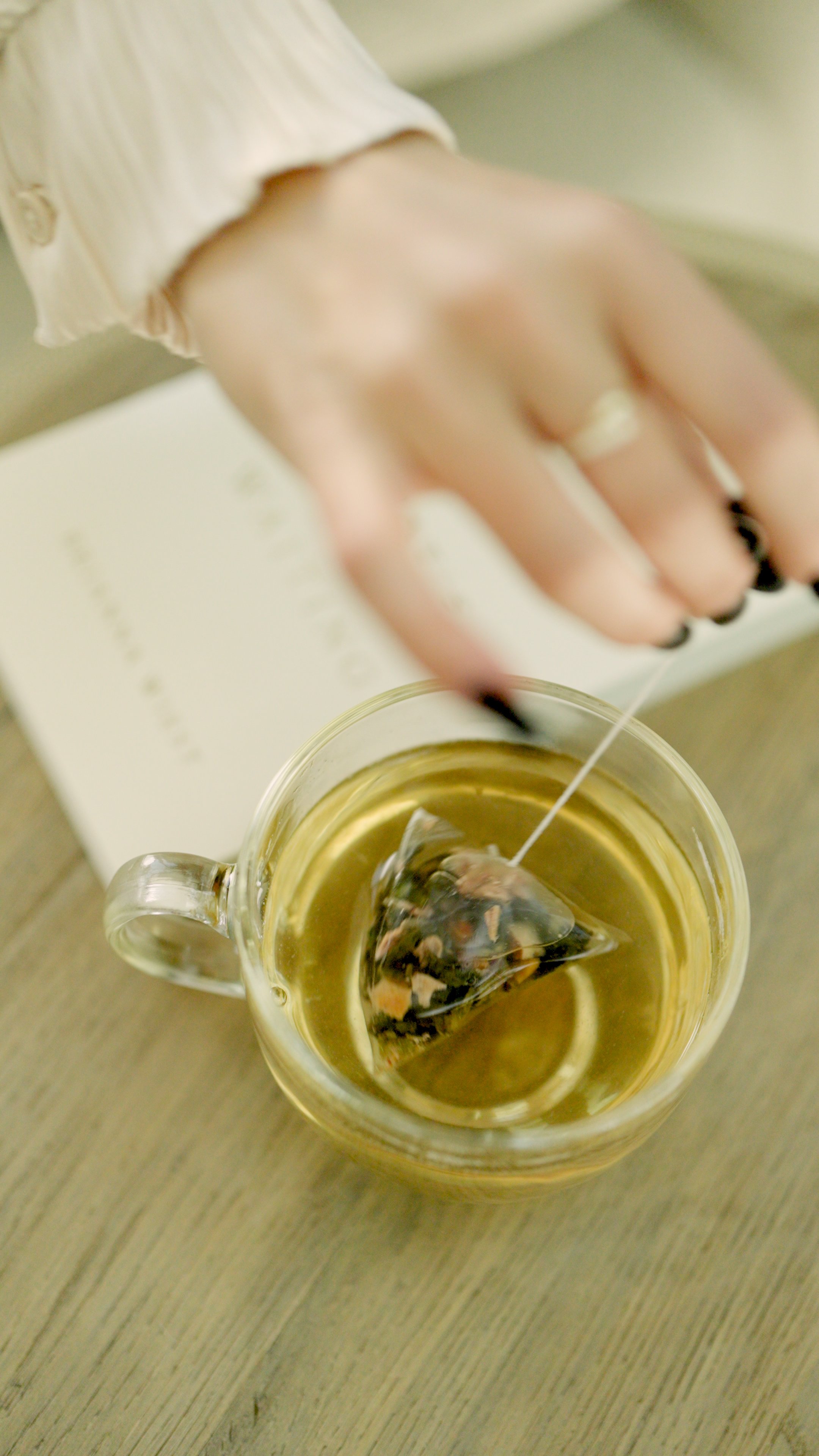 A hand with black painted nails holds a string tea infuser over a glass mug filled with tea, on a wooden surface.
