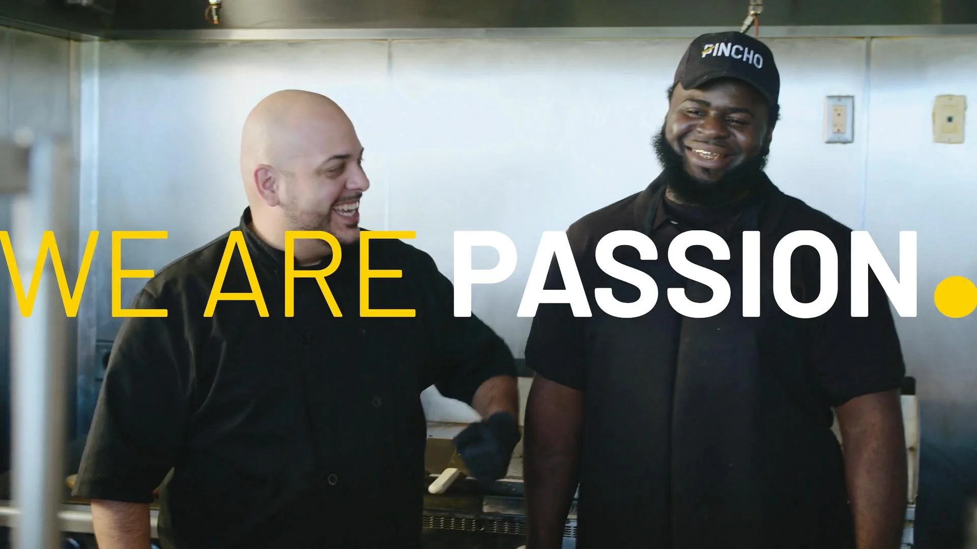 Two smiling male chefs in black uniforms, one wearing a black cap labeled 'PINCHO', standing in a commercial kitchen, with the overlay text 'WE ARE PASSION' in yellow and white letters.