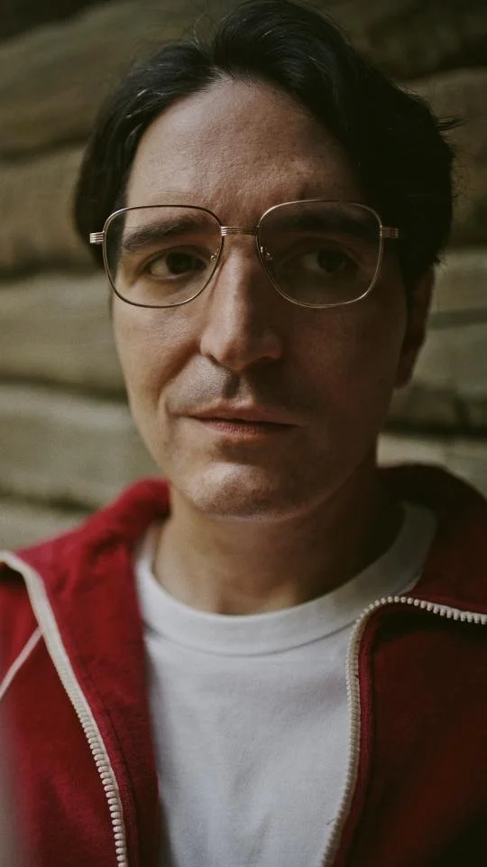 Close-up of a man with dark hair, wearing glasses, a white t-shirt, and a red jacket, standing in front of a wooden background.