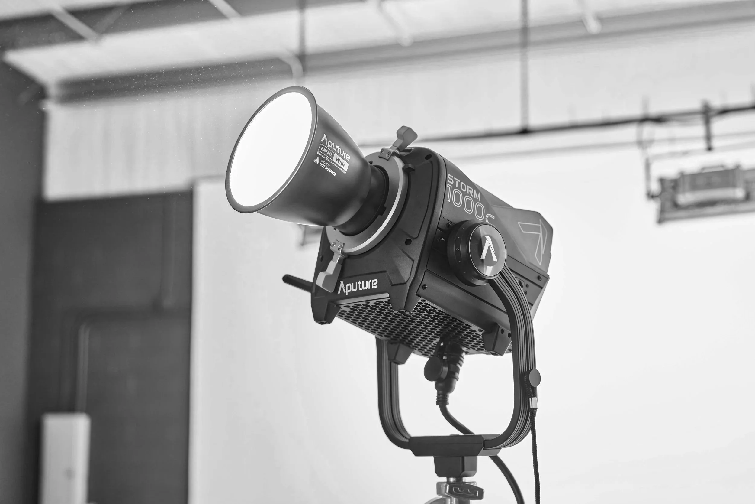 A black and white photograph of an Aputure Storm 1000c LED light on a studio stand, facing upward. The studio environment includes ceiling pipes and some blurred equipment in the background.