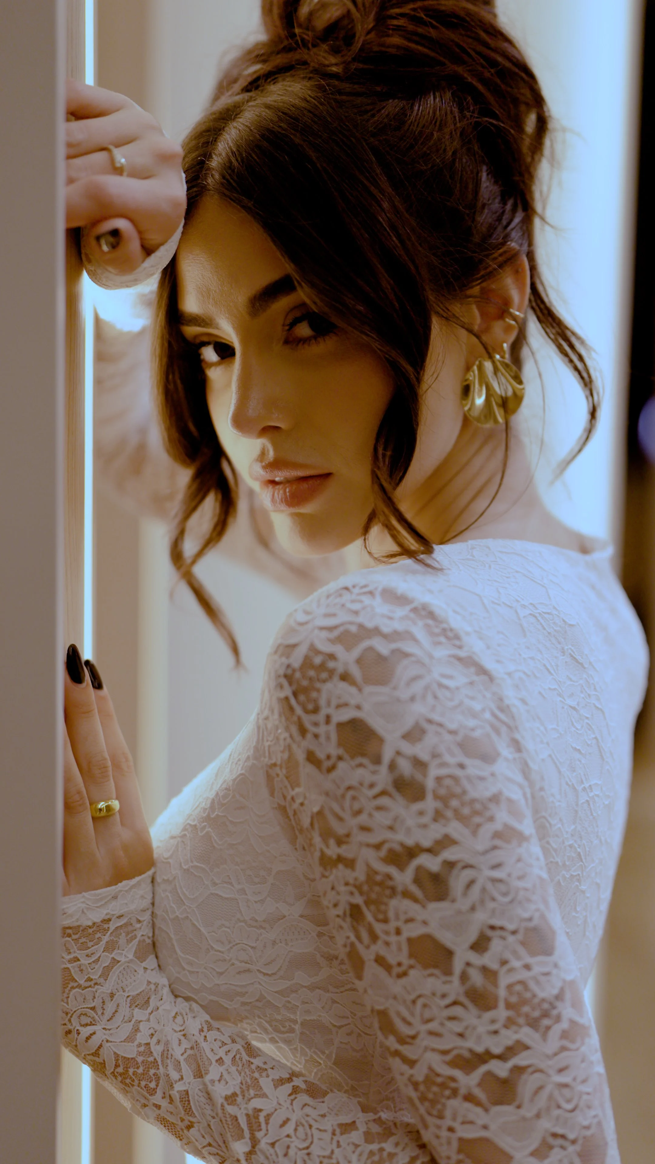A woman with dark wavy hair wearing a white lace dress and gold earrings, looking over her shoulder while standing near a door or wall.