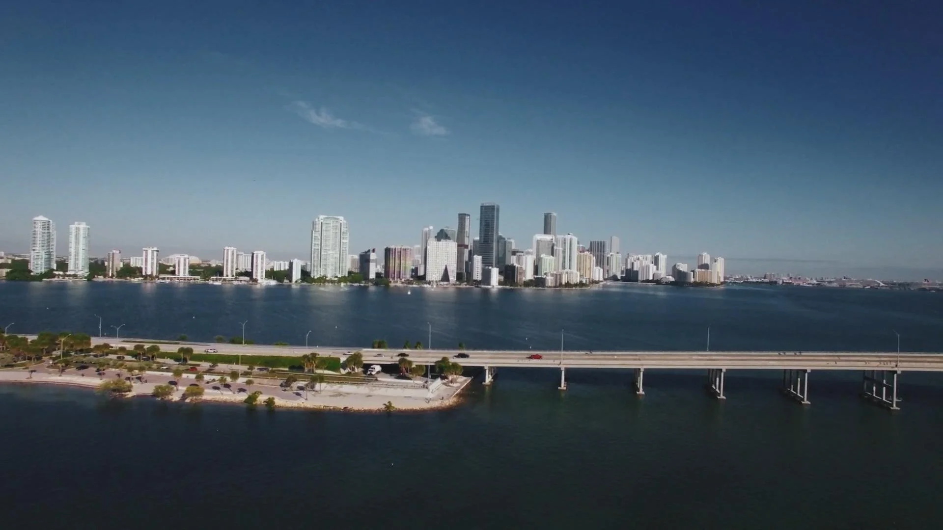 A city skyline with tall modern buildings across a body of water, featuring a bridge and a park with trees in the foreground, under a clear blue sky.