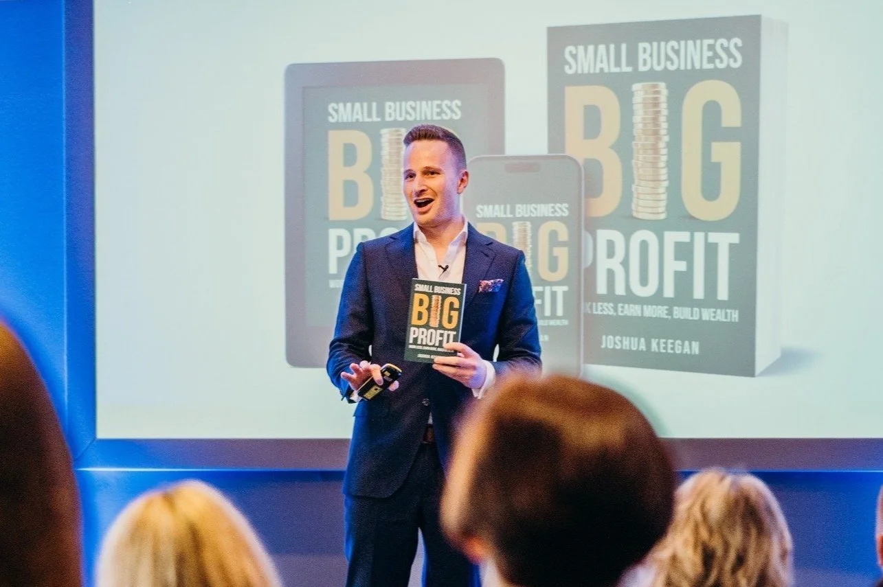 Joshua Keegan in a suit giving a presentation about a book titled 'Small Business Big Profit' to an audience, with a large screen displaying the book cover behind him.