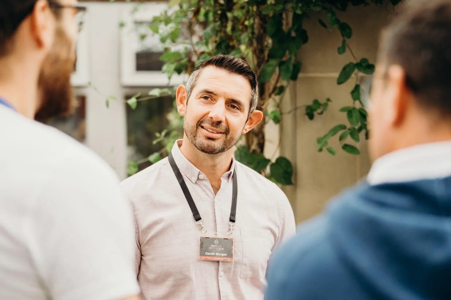 Gareth Morgan with short dark hair and a beard, wearing a light-colored shirt and a nametag, happily talking to two other people outdoors, with greenery and a building in the background.