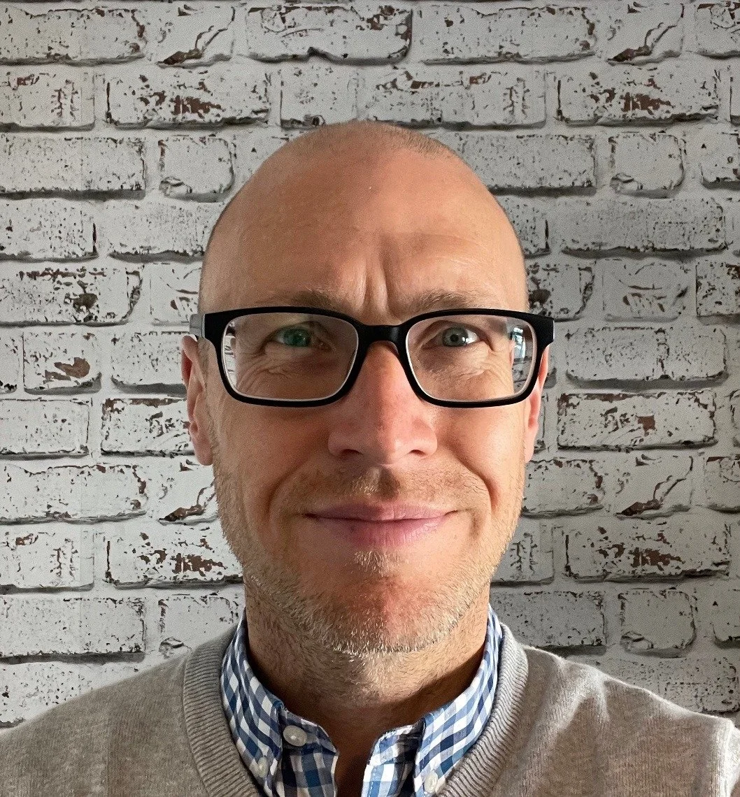 Close-up portrait of a smiling David (man) with glasses, wearing a gray sweater over a checkered shirt, standing in front of a white brick wall.