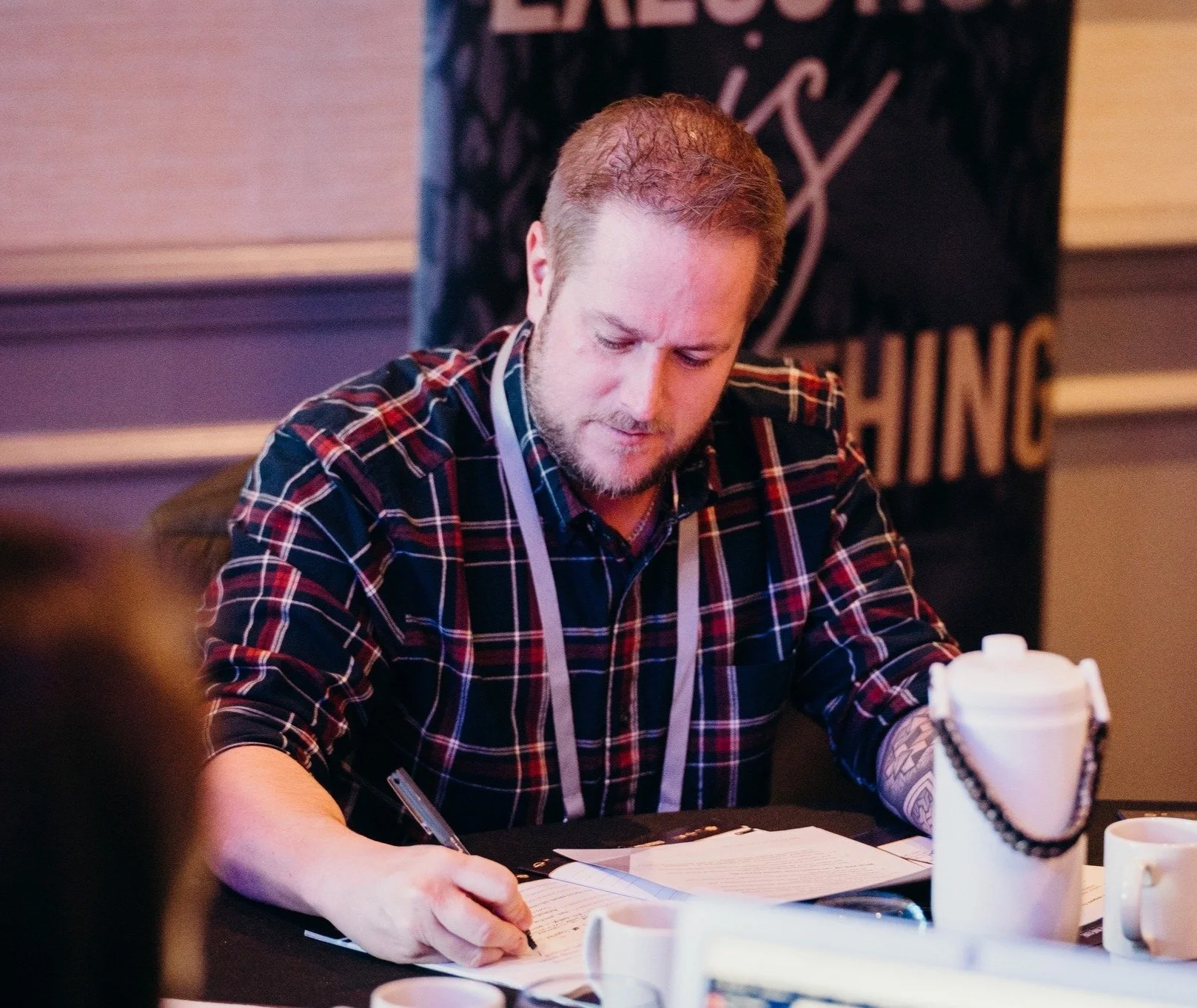 Chris Chadwick with a beard and short hair, wearing a plaid shirt, is sitting at a table writing on paper with a pen. There are cups and a white insulated container on the table. In the background, there is a black sign with partially visible text.