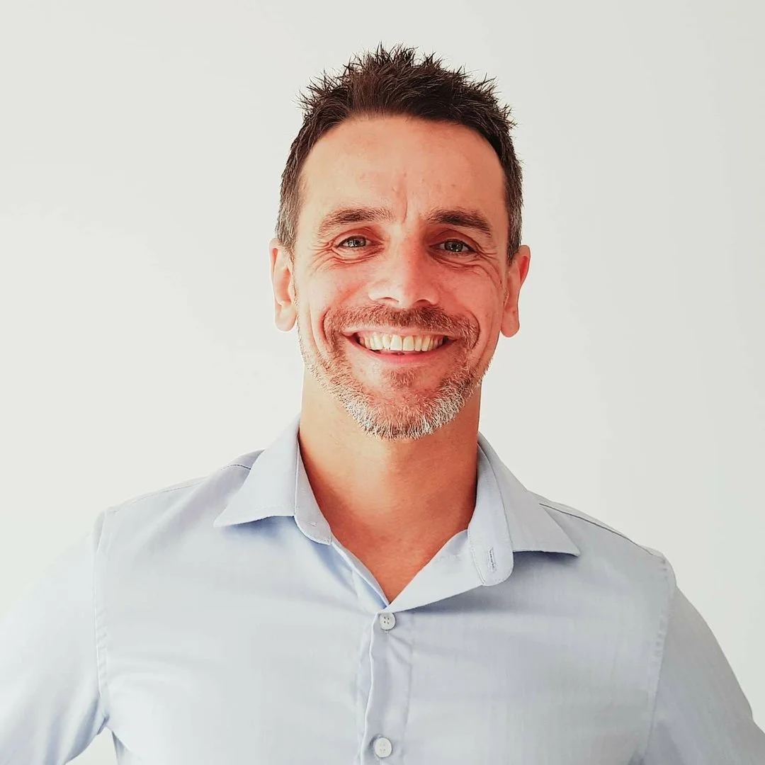 A smiling man with short dark hair, blue eyes, and facial hair, wearing a light blue dress shirt, standing against a plain white background.