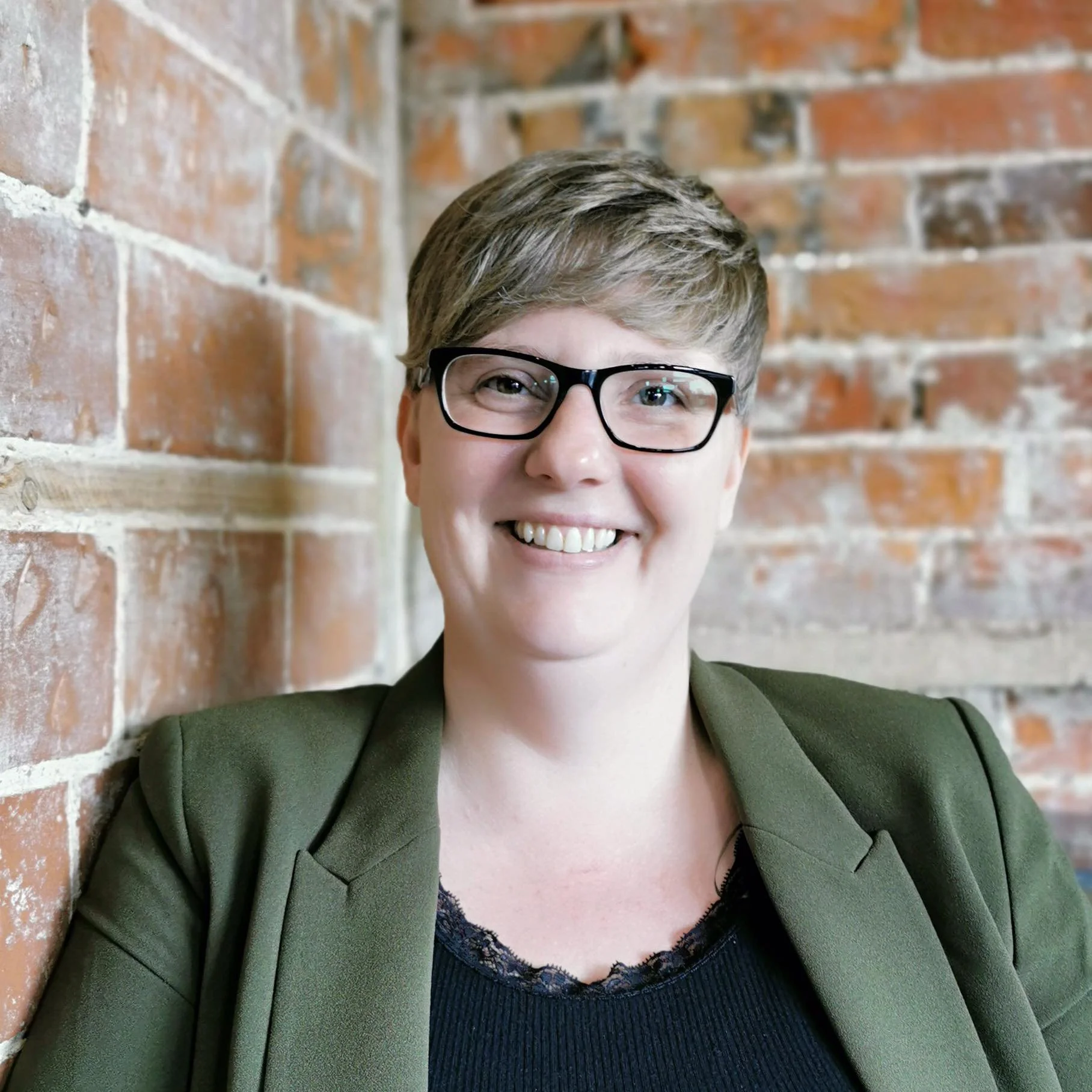 A woman with short brown hair, glasses, and a smile, wearing a green blazer over a black top, posing against a brick wall.
