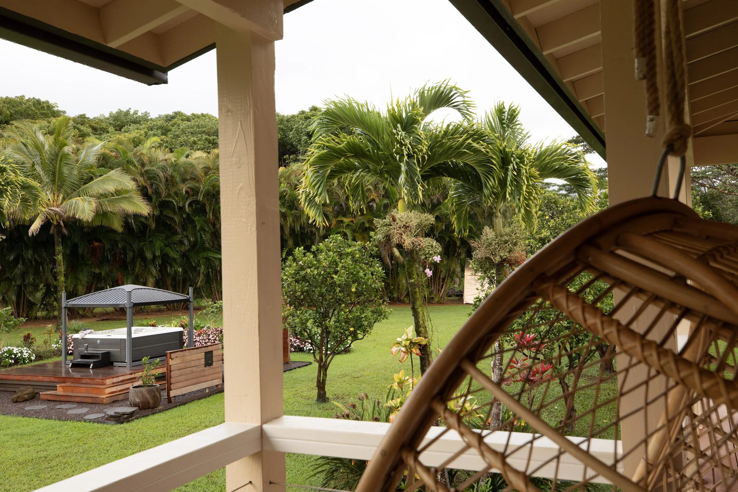 View from a porch showing lush tropical garden with palm trees, small flowering shrubs, a hot tub with a roof cover, and a wooden deck area, with a wicker chair in the foreground.