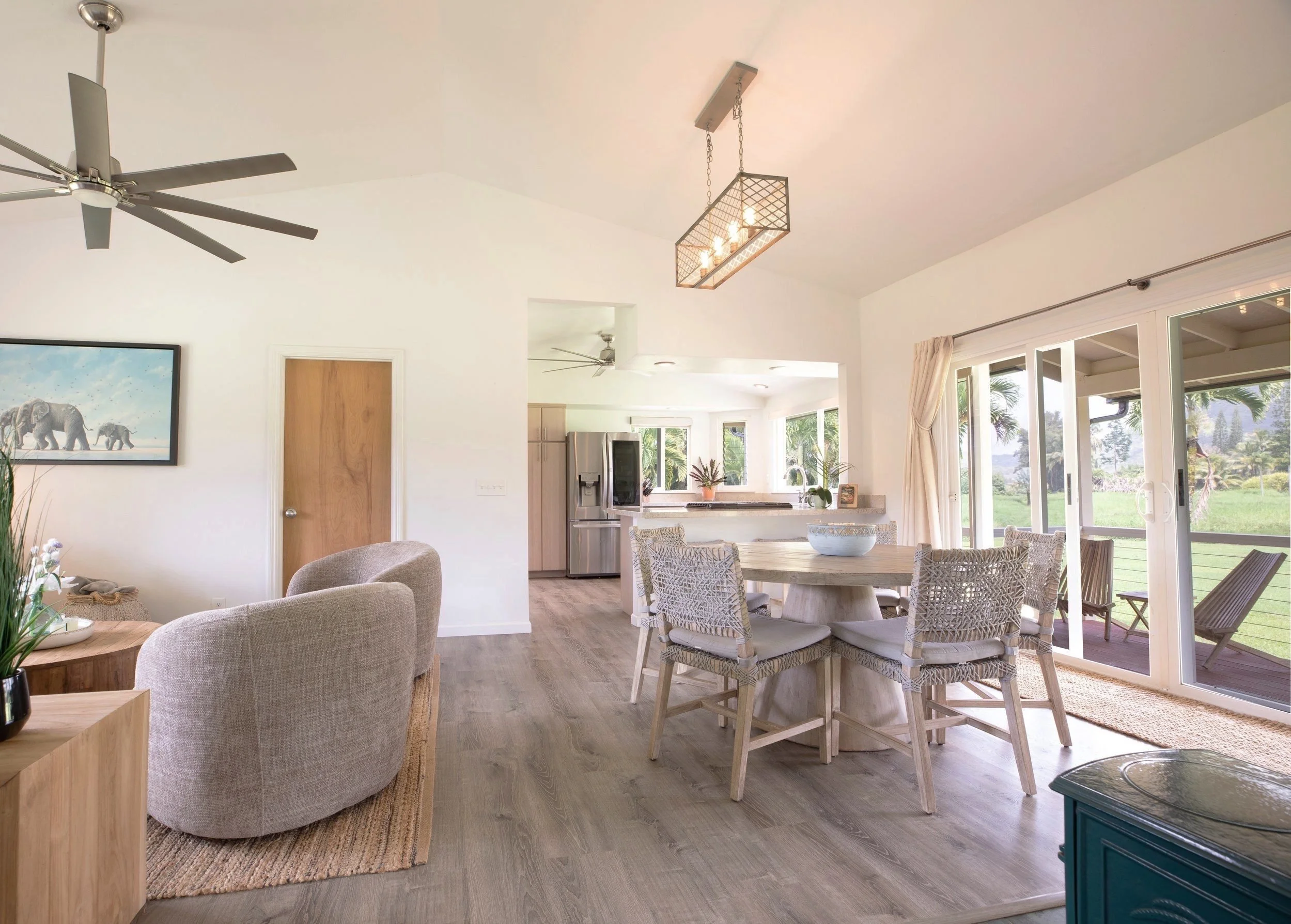 Open-concept living and dining area with natural light, wooden flooring, wicker and upholstered furniture, modern ceiling fan, and large sliding glass doors leading to a porch with outdoor seating, overlooking a green landscape.