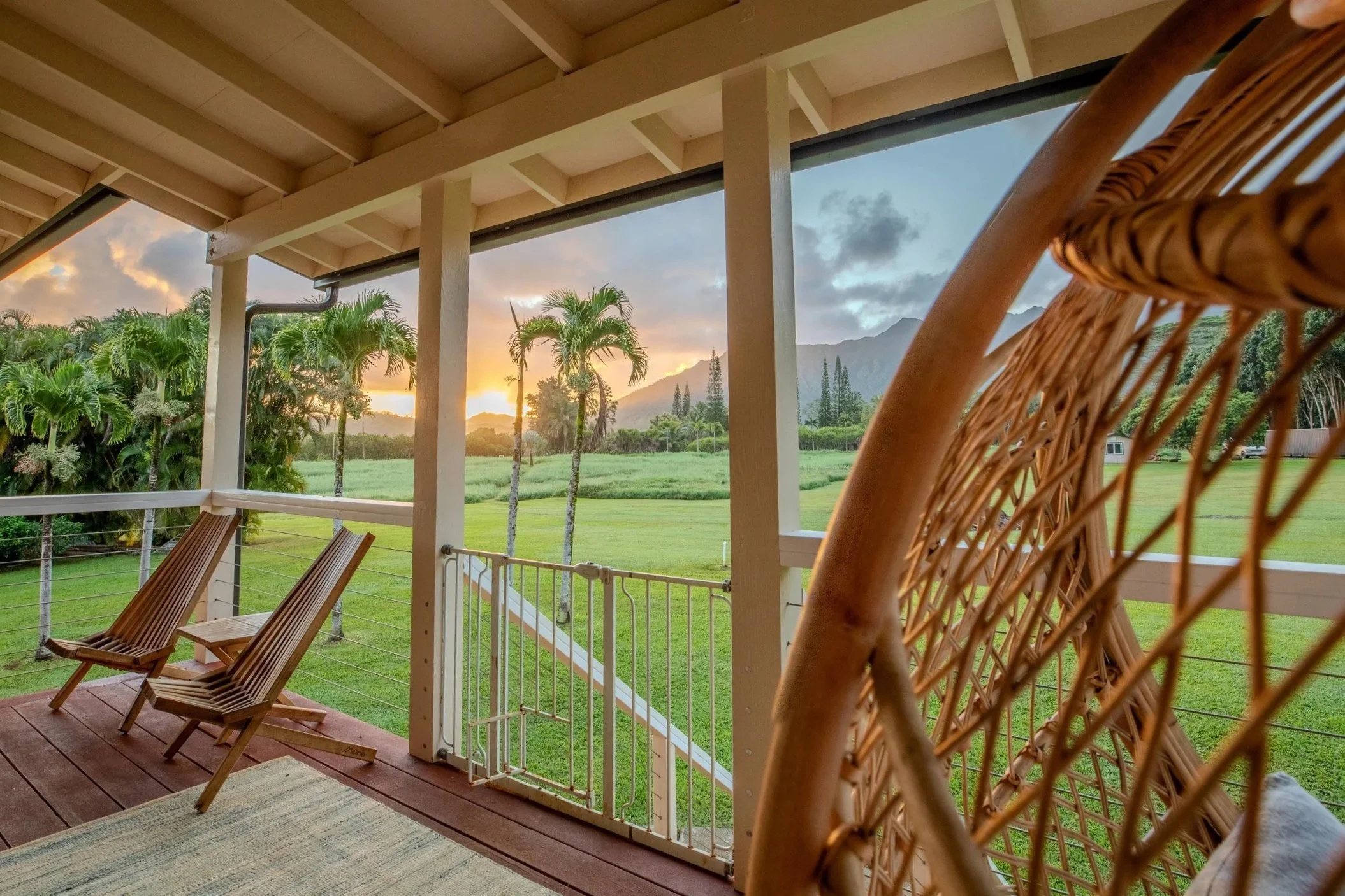 Sunset view from a porch with wooden chairs and a wicker chair, overlooking a lush green field with palm trees and mountains in the background.