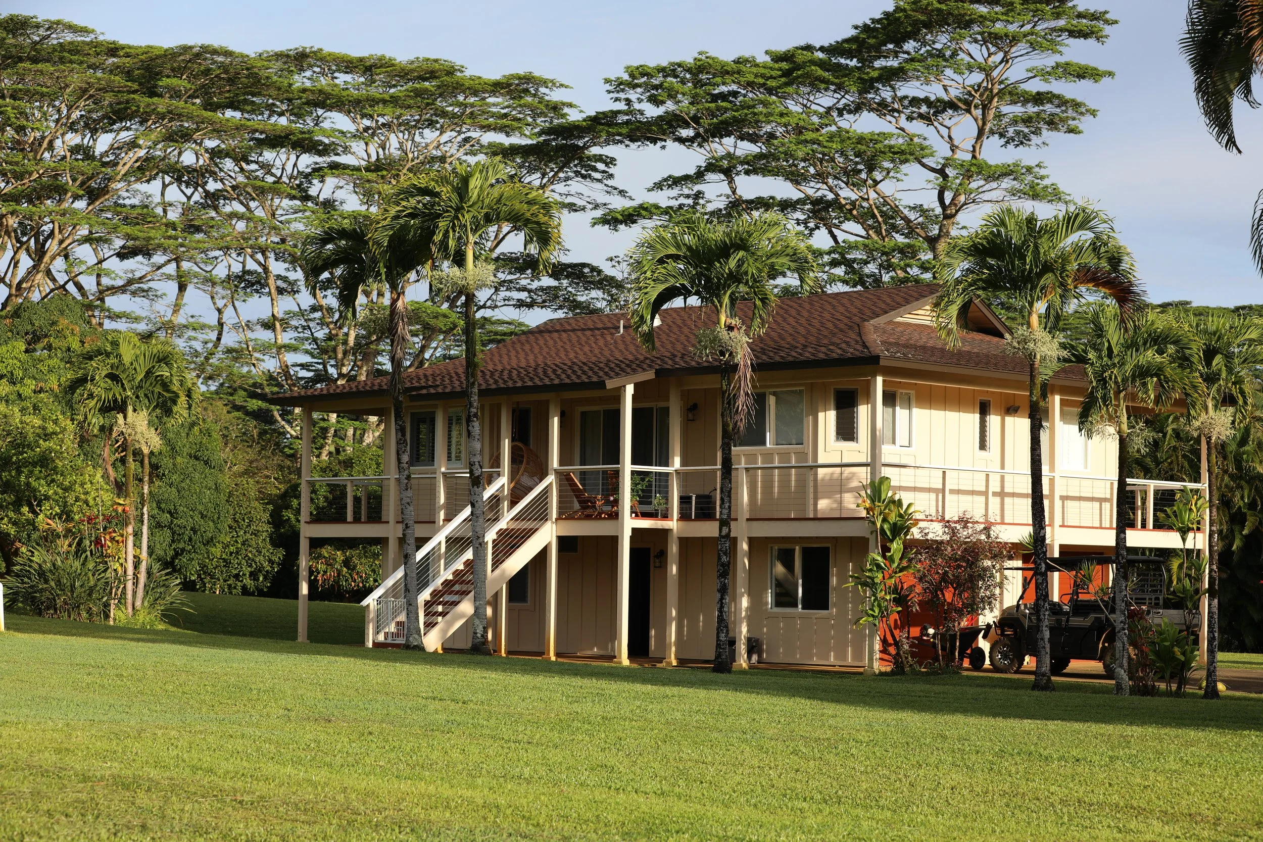A two-story house with a brown roof surrounded by tall trees and palm trees in a grassy yard.