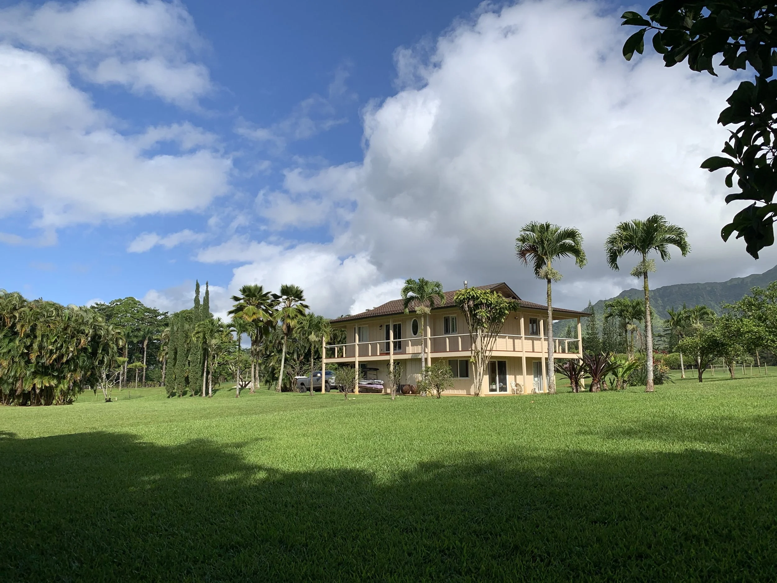 A two-story house with beige exterior and a red roof, surrounded by tall palm trees, lush green grass, and tropical plants, under a partly cloudy sky with mountains in the background.