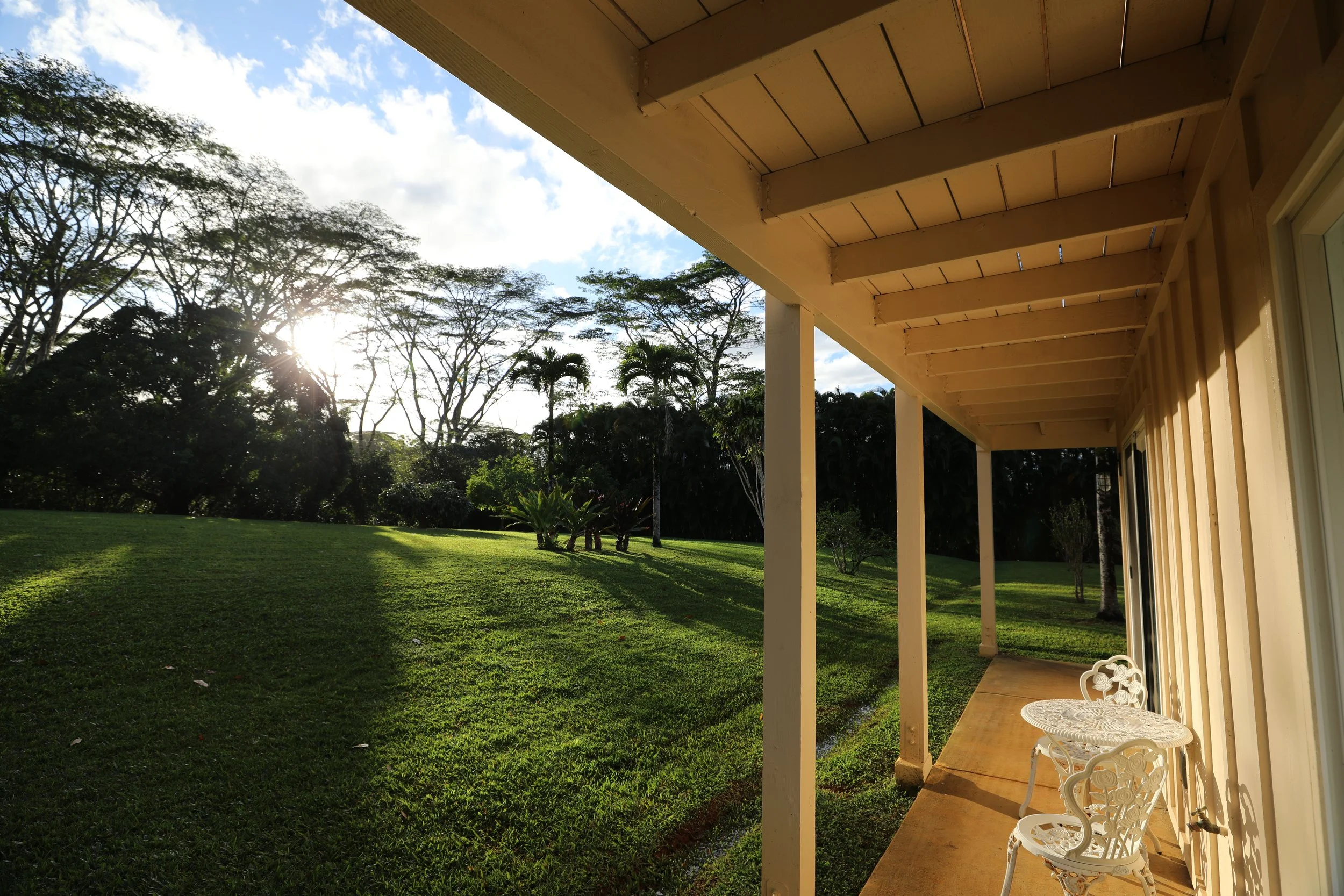 Sunset view from a porch with a small white table and chairs, overlooking a lush green yard and trees.