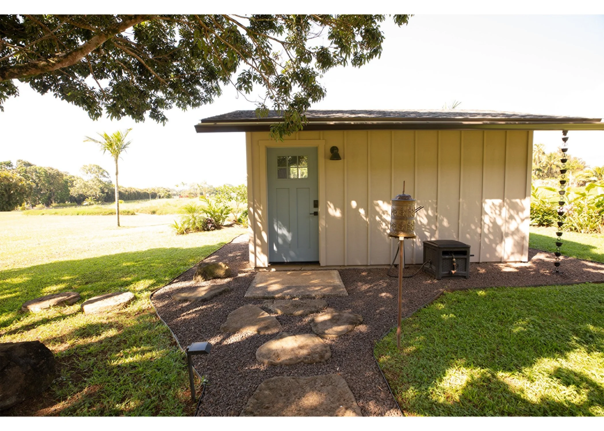 Small beige guest cottage/office with light blue door, surrounded by a landscaped yard with a stepping stone pathway, trees, and grass.