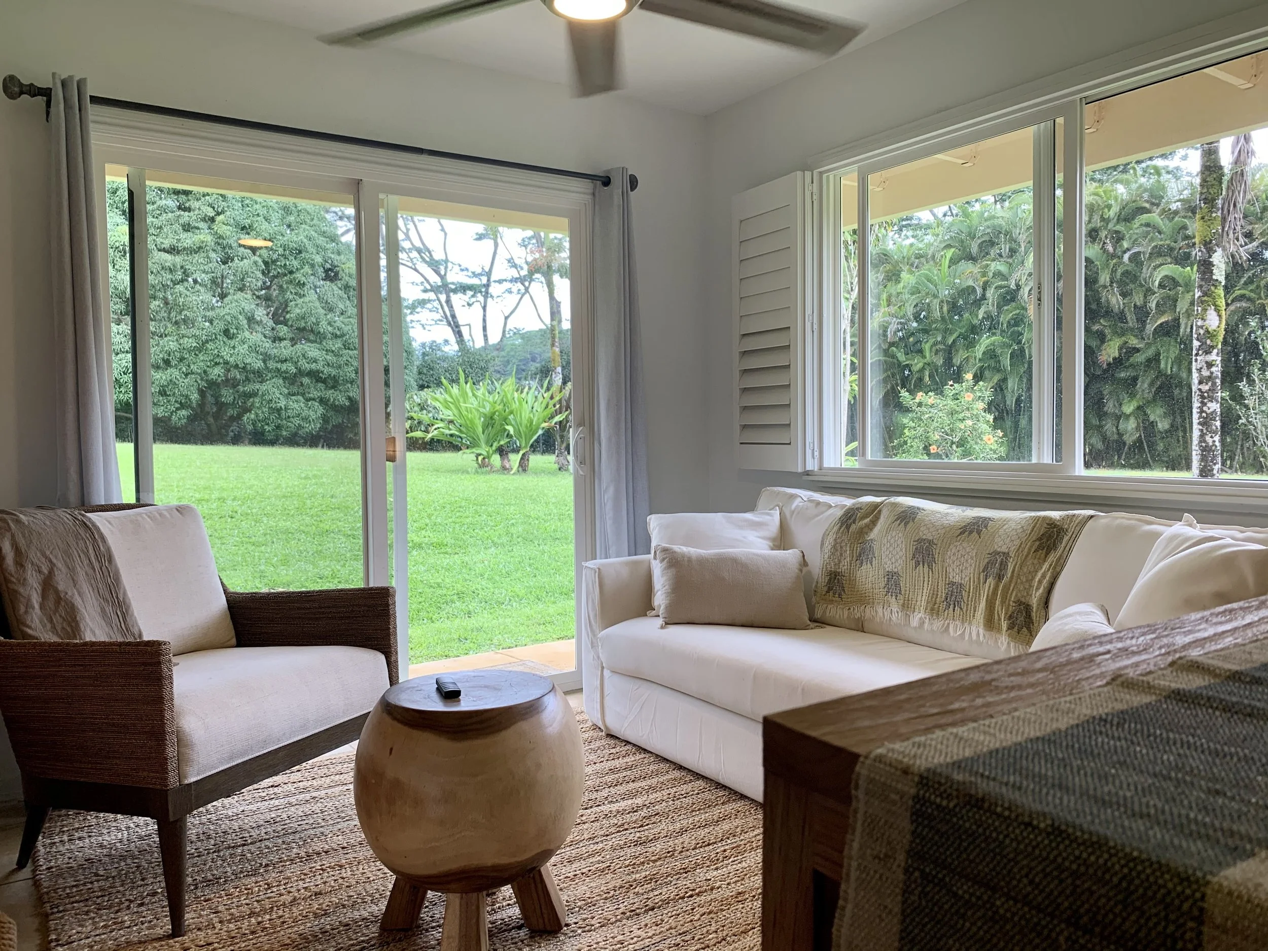 Living room with a white sofa, beige cushions, a patterned blanket, a beige armchair, a small wooden stool, and large windows showing a lush green garden outside.