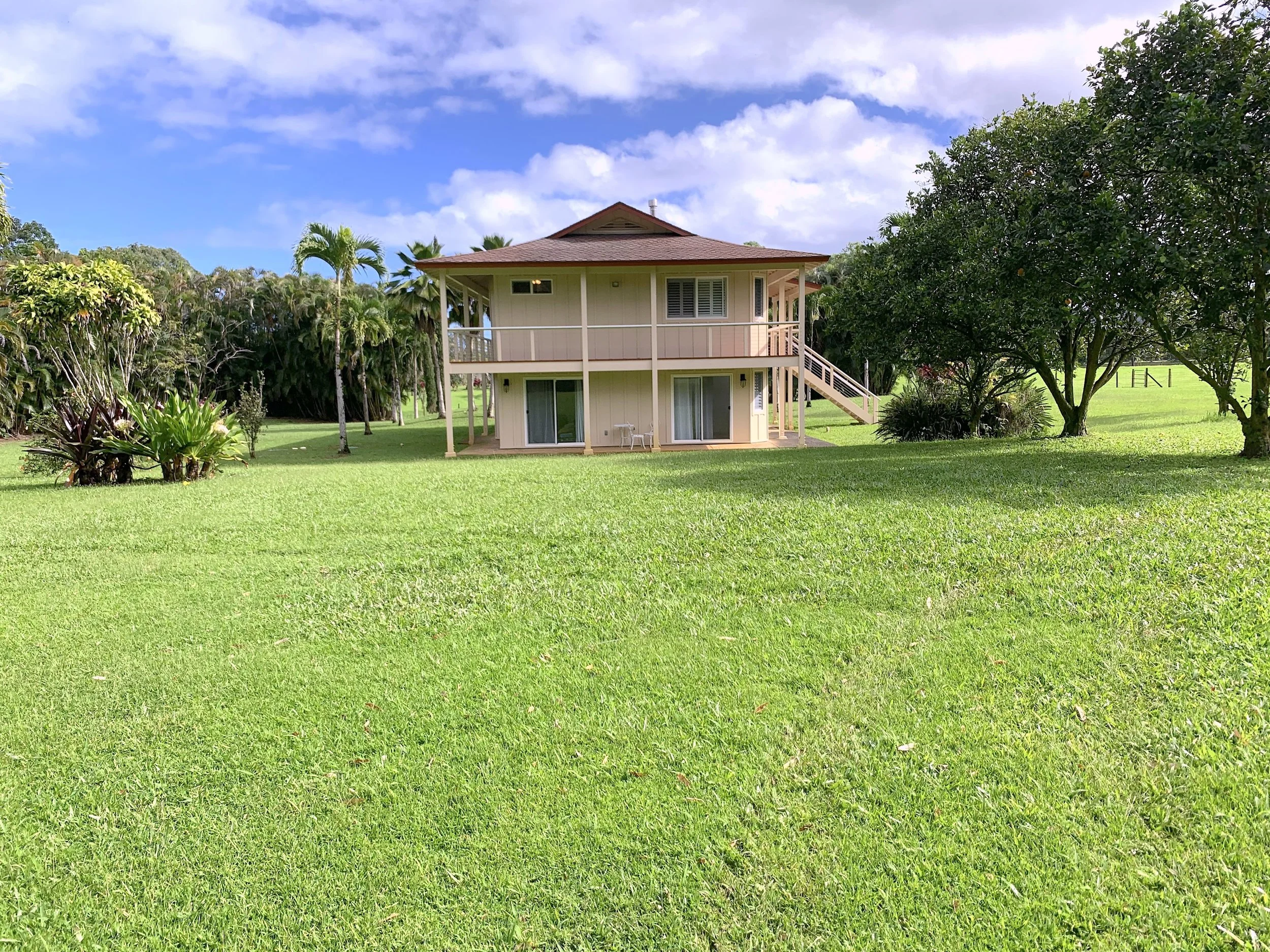 A two-story house with a porch and staircase, surrounded by green grass, trees, and bushes under a partly cloudy sky.