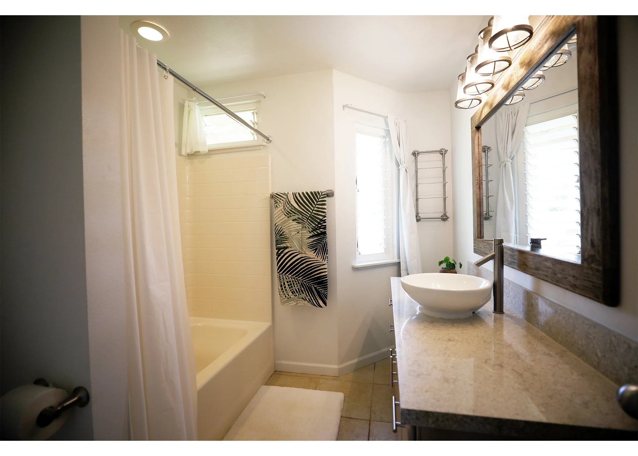 A modern bathroom with a vanity mirror, vessel sink, granite countertop, a window with white shutters, a towel rack, and a bathtub with a shower curtain.