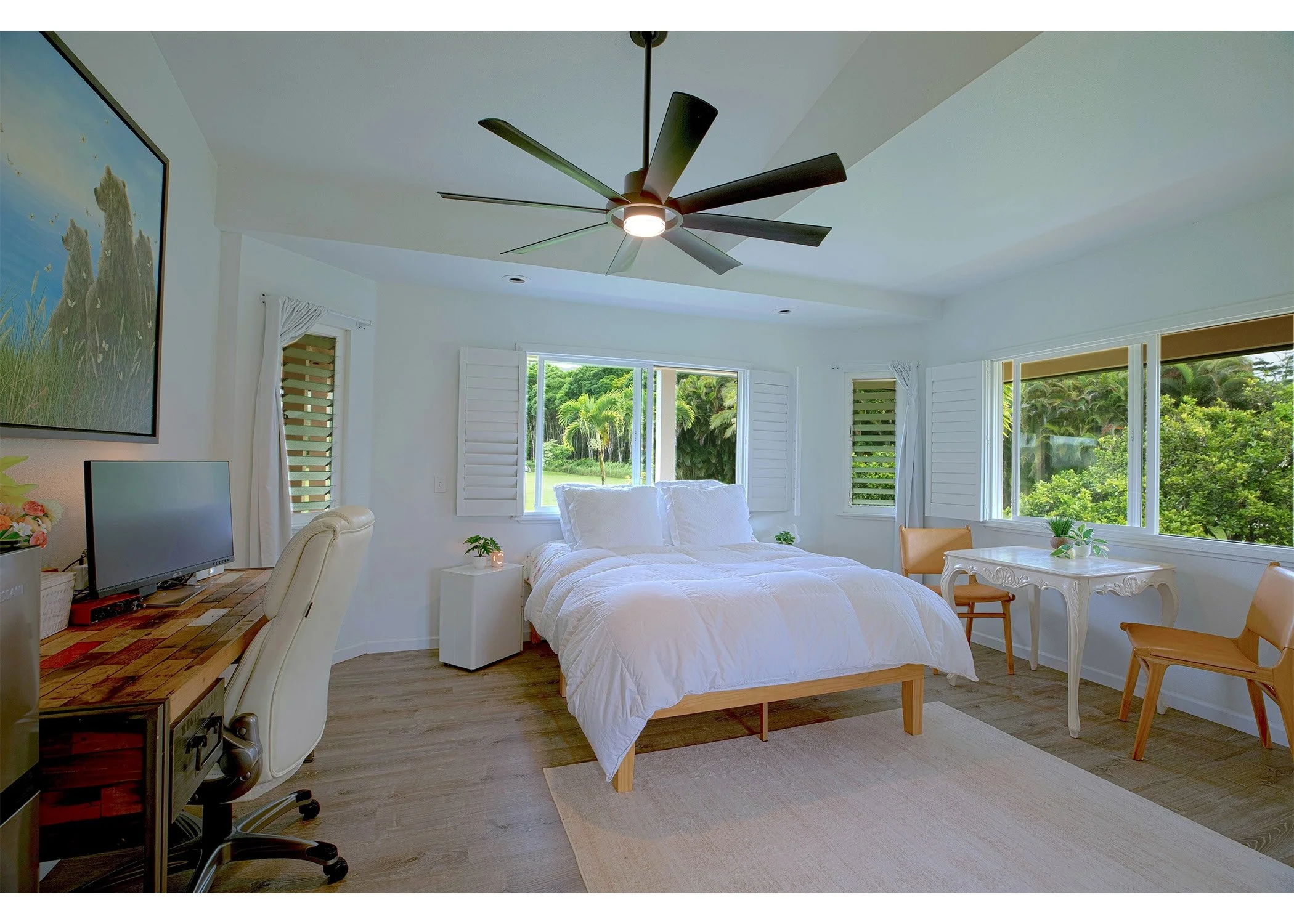 Bright bedroom with large windows showing lush greenery outside, white bed with pillows, ceiling fan, small desk with computer, and a white decorative table with chairs.