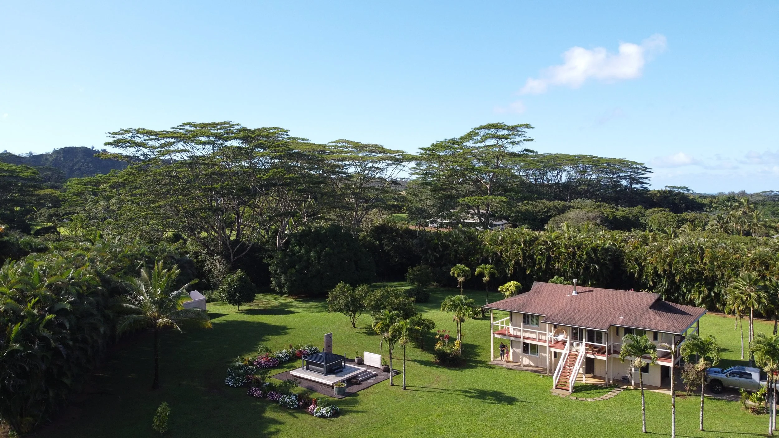 A large house with a brown roof and a white exterior, surrounded by lush green grass, palm trees, and colorful flower gardens, under a clear blue sky.