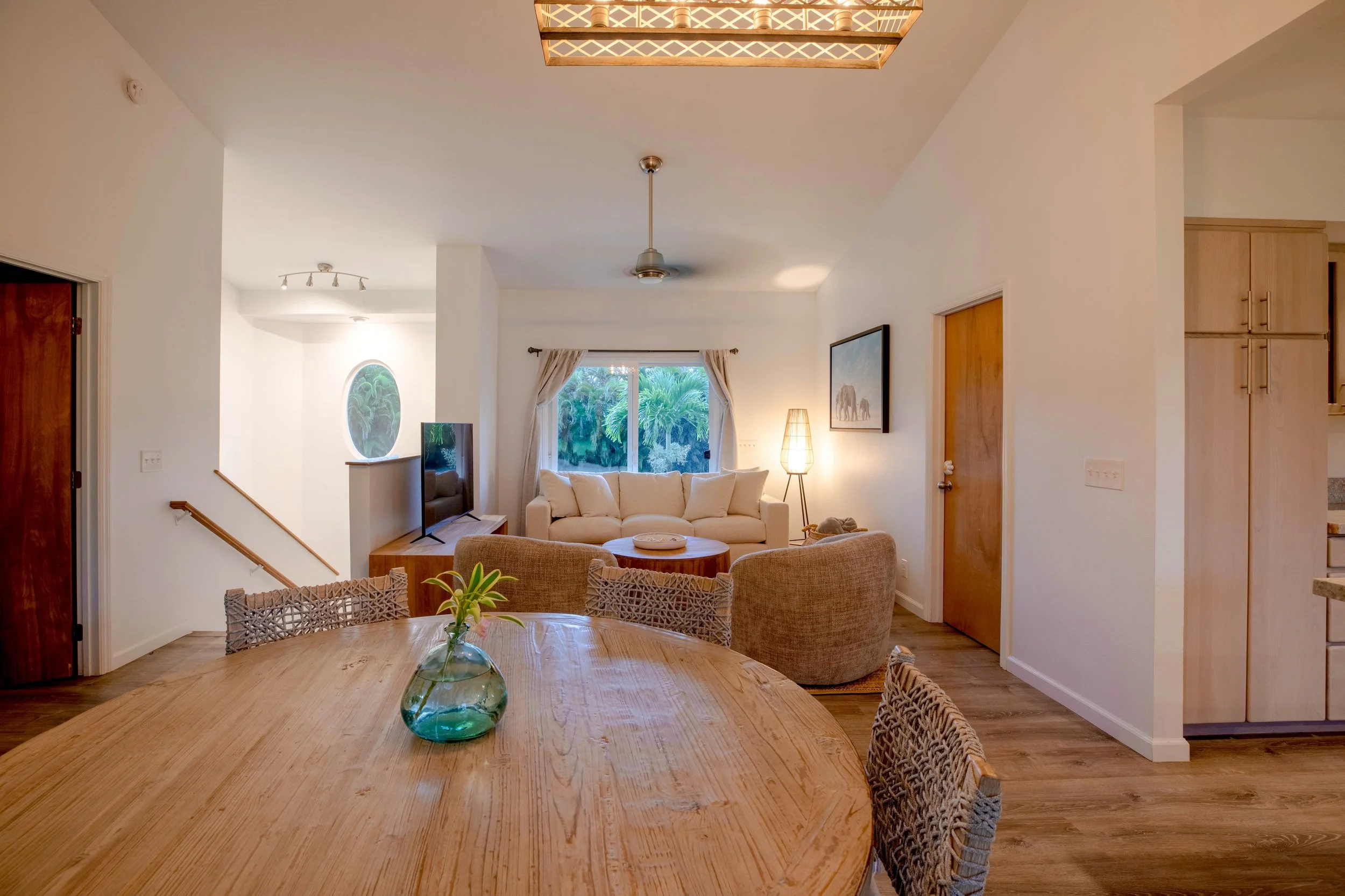 Modern living and dining room with wooden furniture, beige sofa, round dining table, woven chairs, floor lamp, TV, and large window.