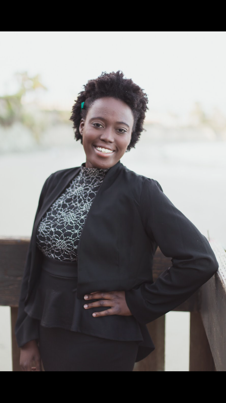 Smiling person standing outdoors with a railing, wearing a black jacket and patterned top.