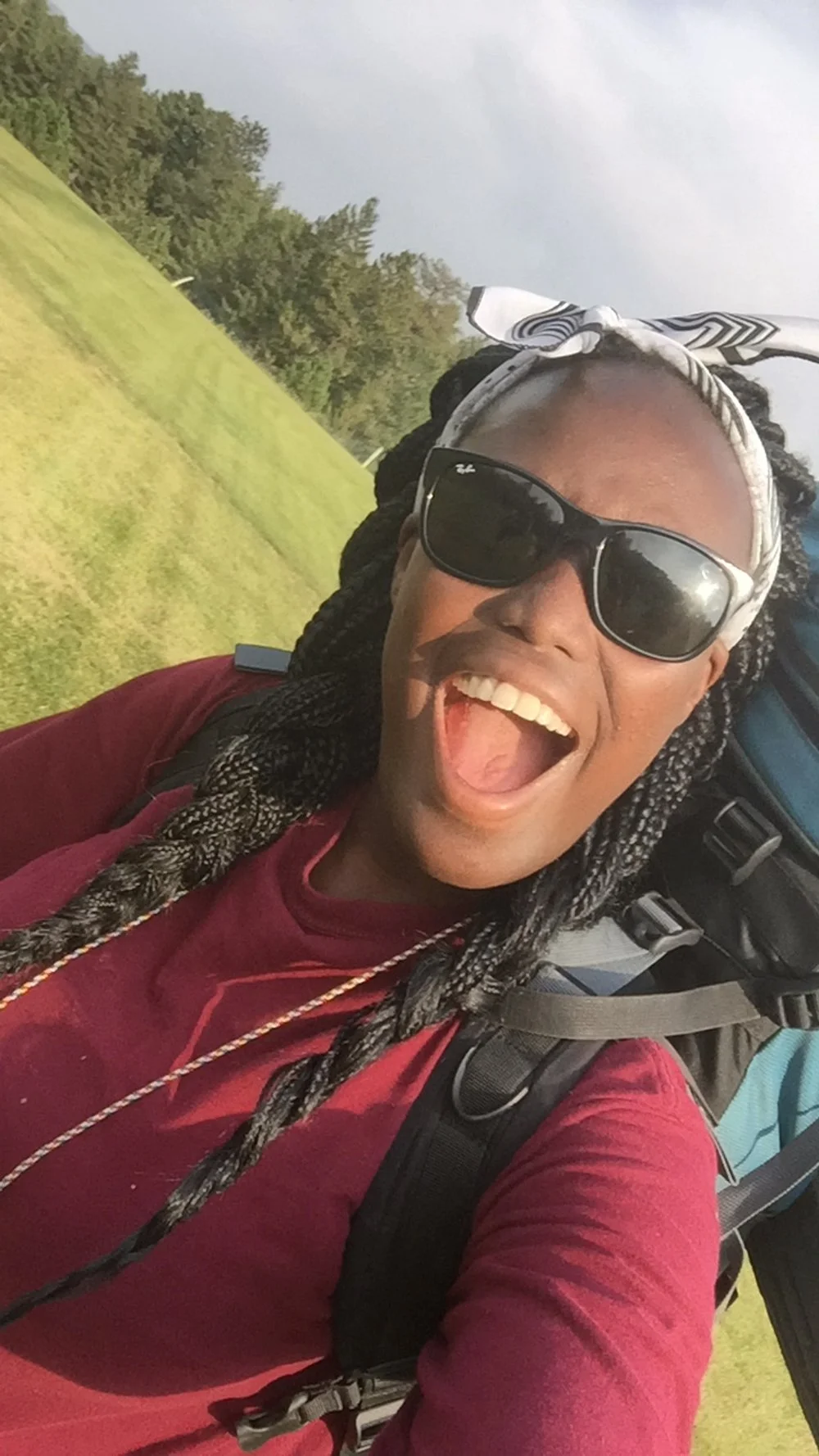 Person with braids and sunglasses smiling with a backpack outdoors.