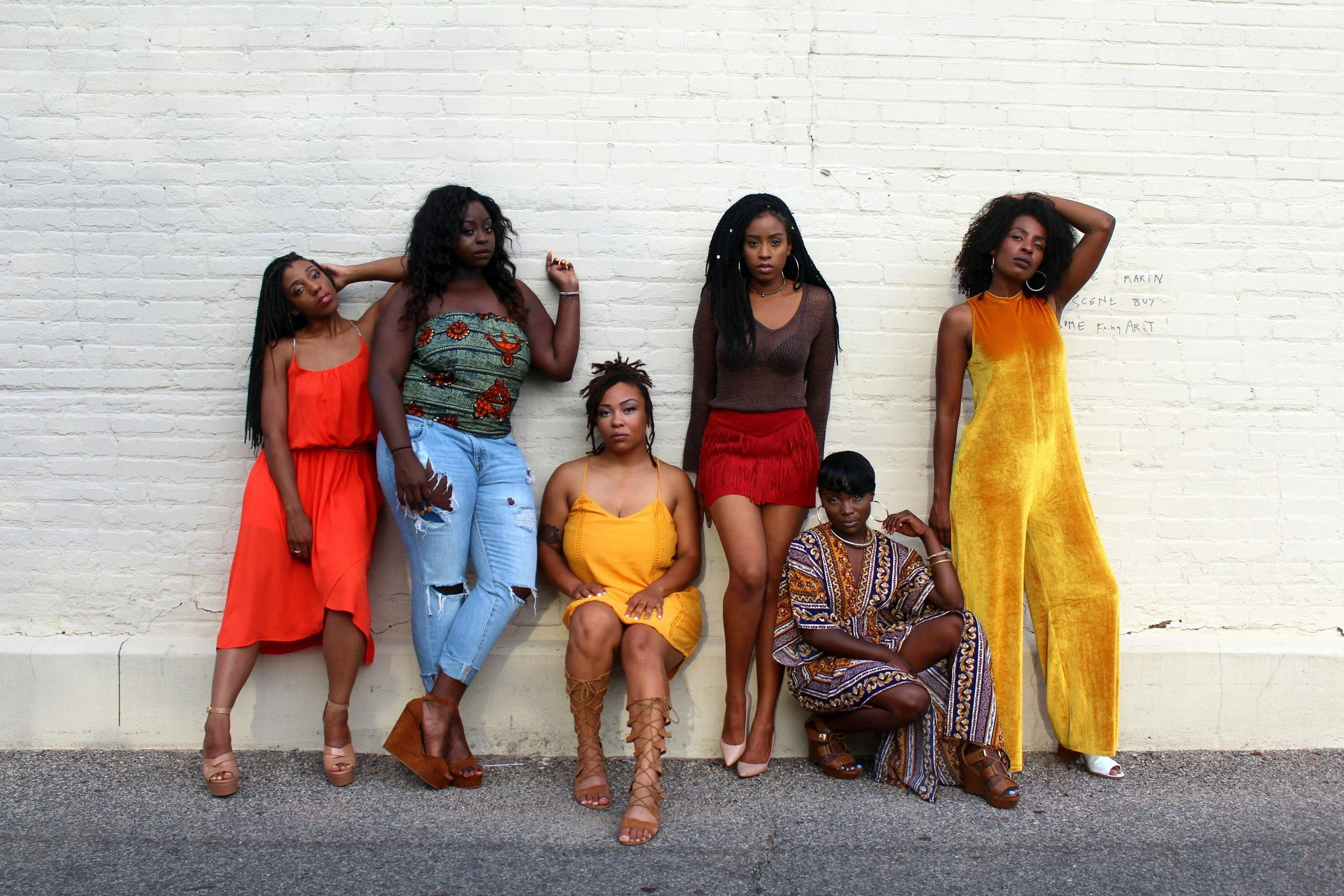 Group of six women posing against a white brick wall in colorful outfits, including dresses and jeans, with diverse hairstyles, showcasing fashion and individuality.
