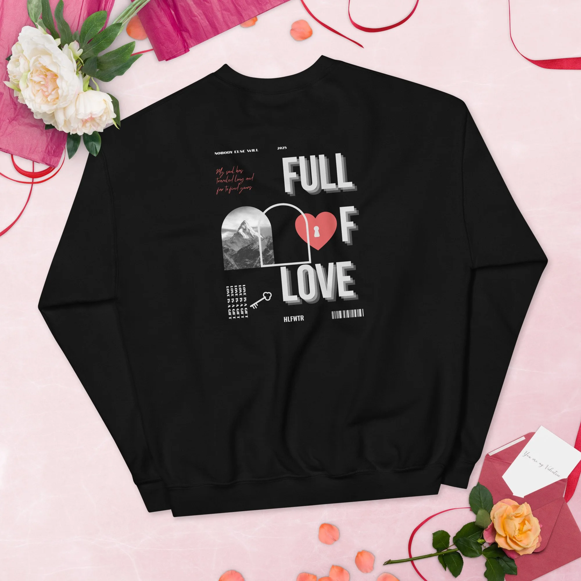 Black sweatshirt with graphic text and design, surrounded by flowers, a pink envelope with a card, and peach-colored rose petals, on a pink background.