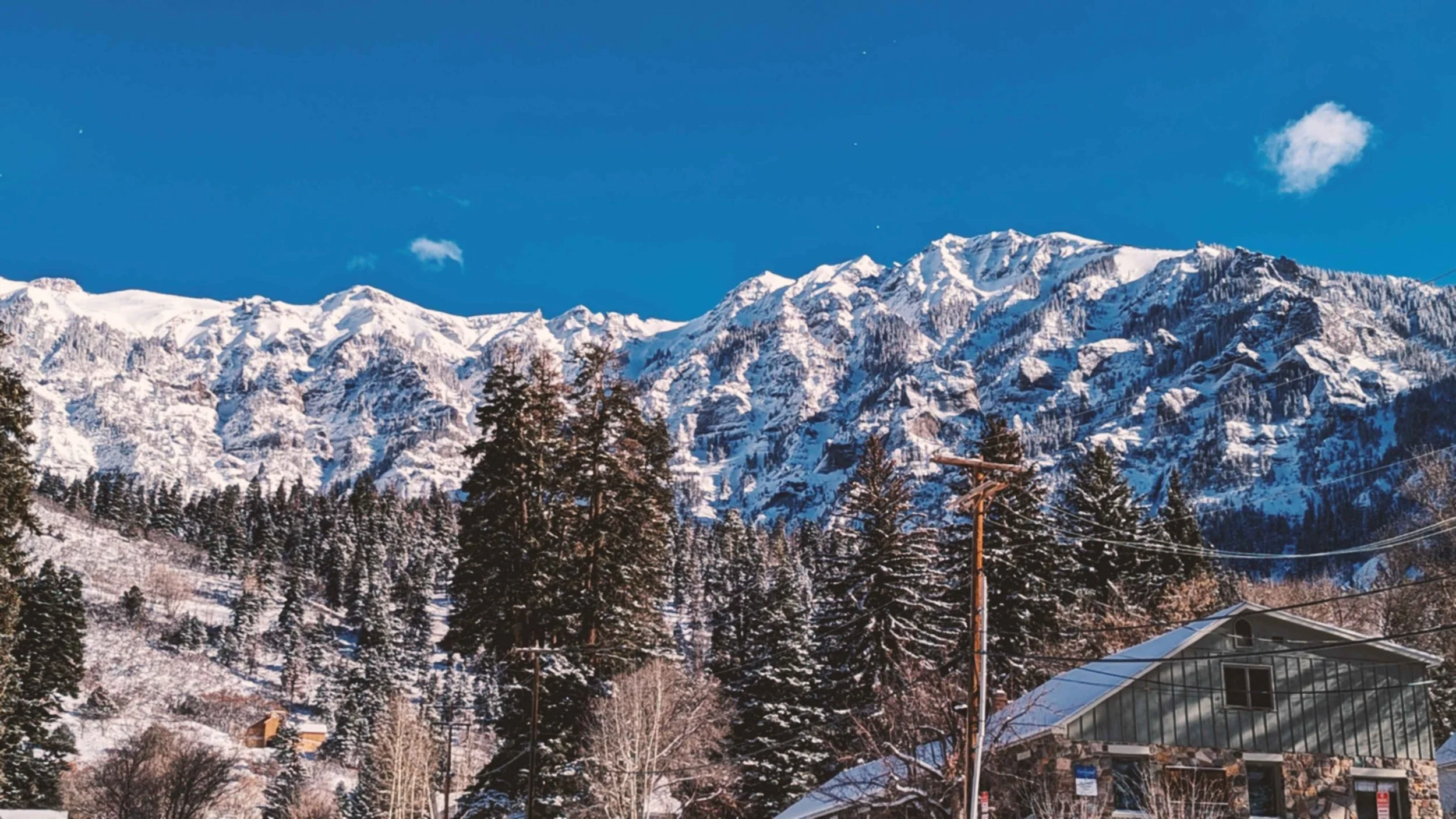 Snow-covered mountain range under a clear blue sky with a few clouds, with snow-topped evergreen trees and a small building with a metal roof in the foreground.