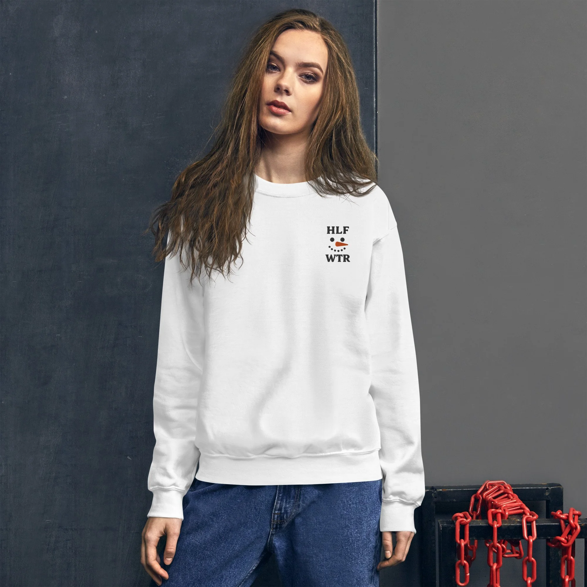 A young woman with long, wavy brown hair wearing a white sweatshirt with small black and orange graphic text and design, standing against a dark background with a red chain accessory on a black box nearby.