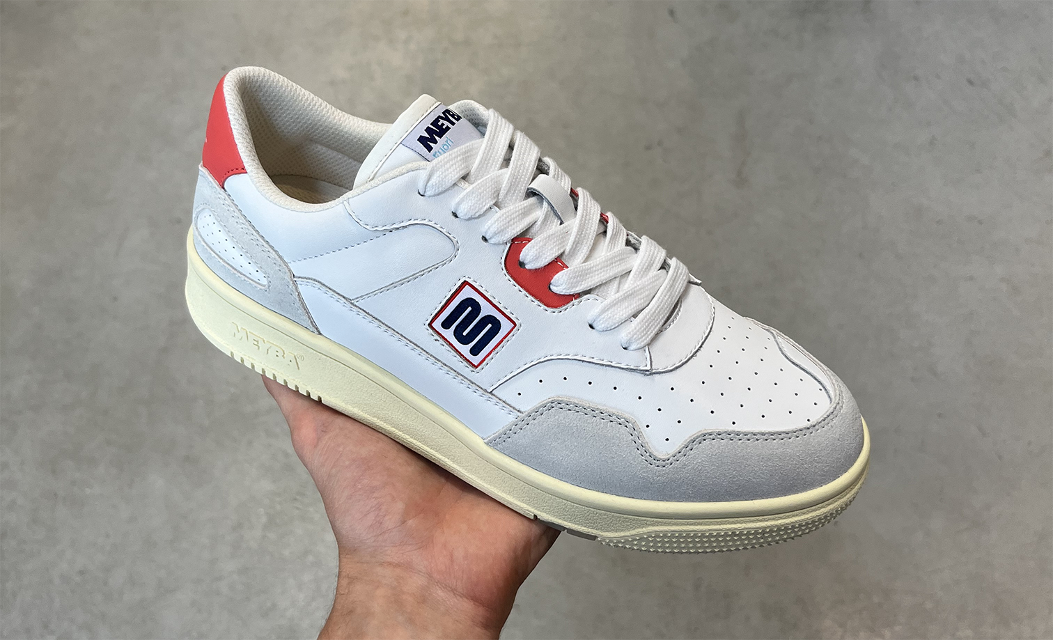 White sneaker with red and blue accents, held in hand against a gray background.